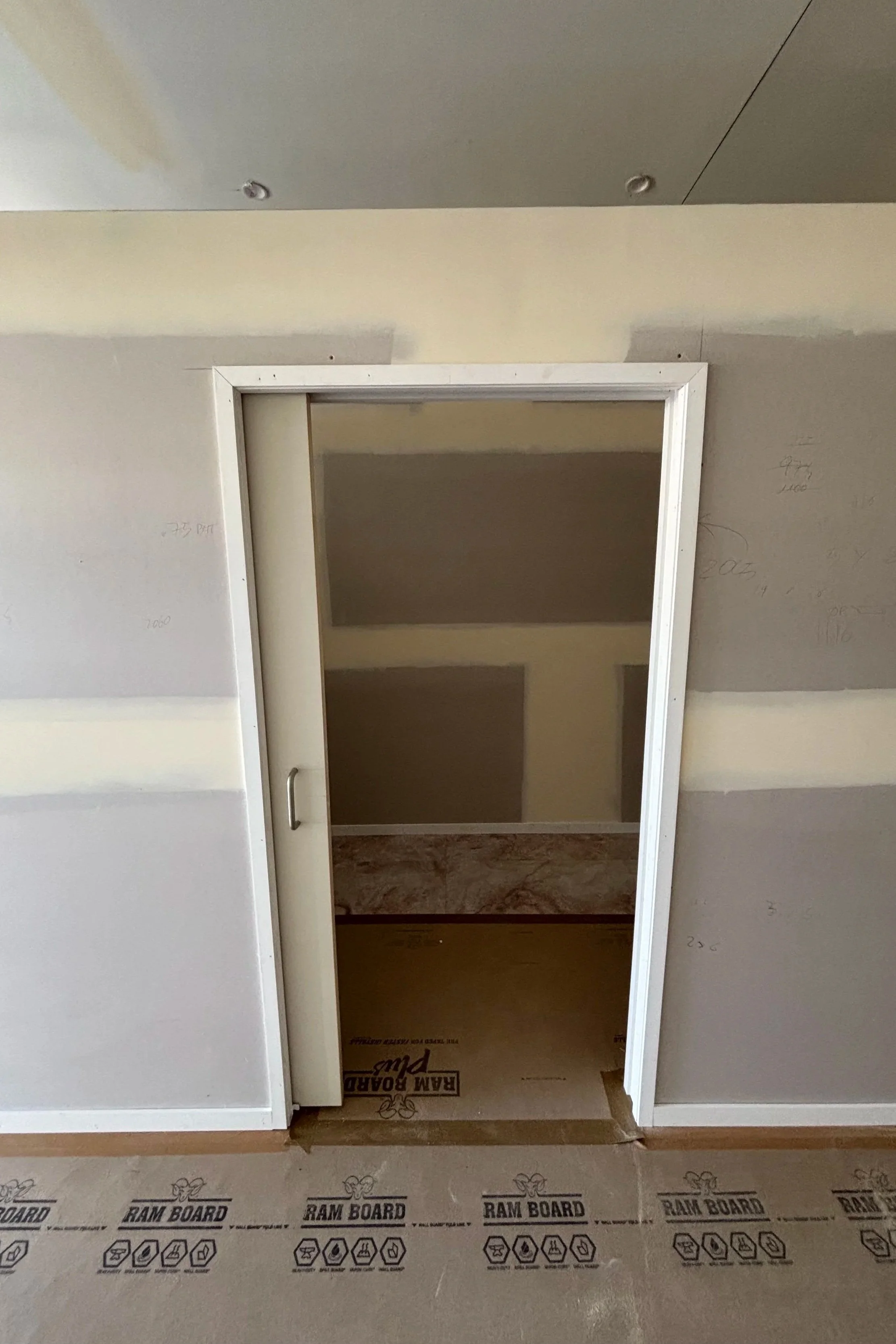 View of an interior doorway with unfinished walls, partially painted, leading to a small room or closet. The floor is covered with RAM BOARD protective material, indicating ongoing construction or renovation.