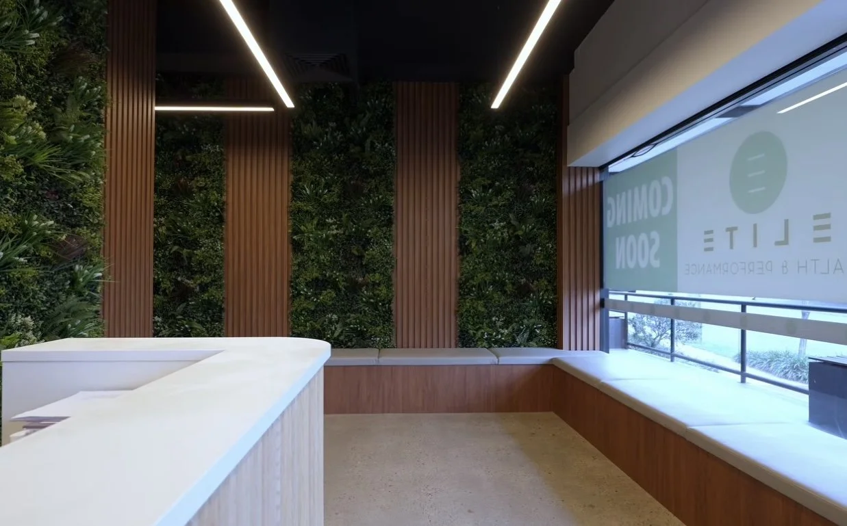 Empty waiting area with a white desk, green plants on the wall, wooden panels, and a large window with a screen.