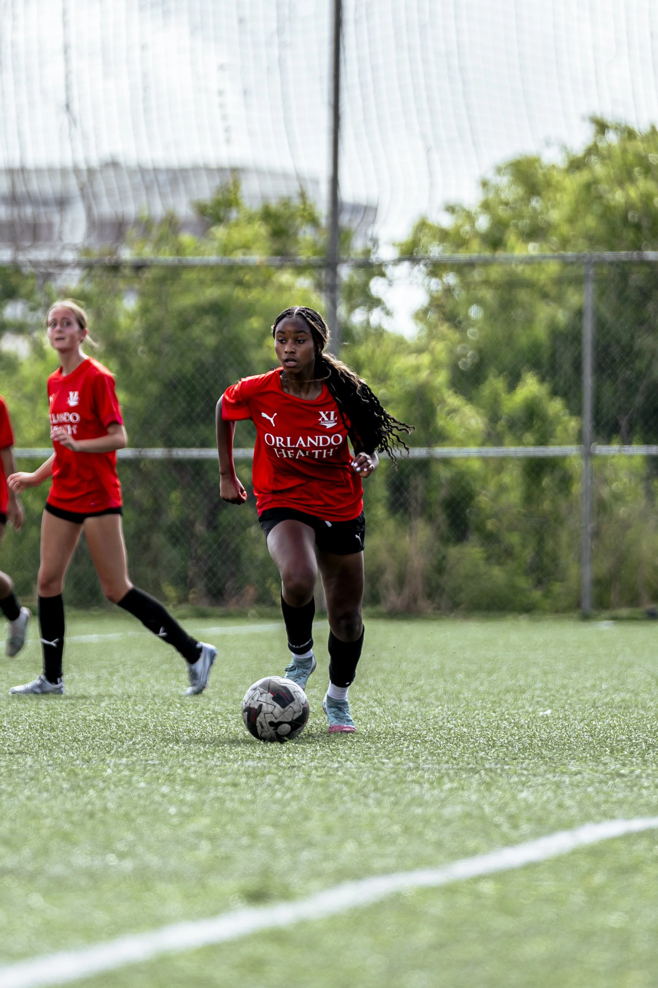 A girl in a red Orlando Health soccer jersey dribbling a soccer ball on a green field during daylight, with other girls in similar jerseys nearby and trees in the background.