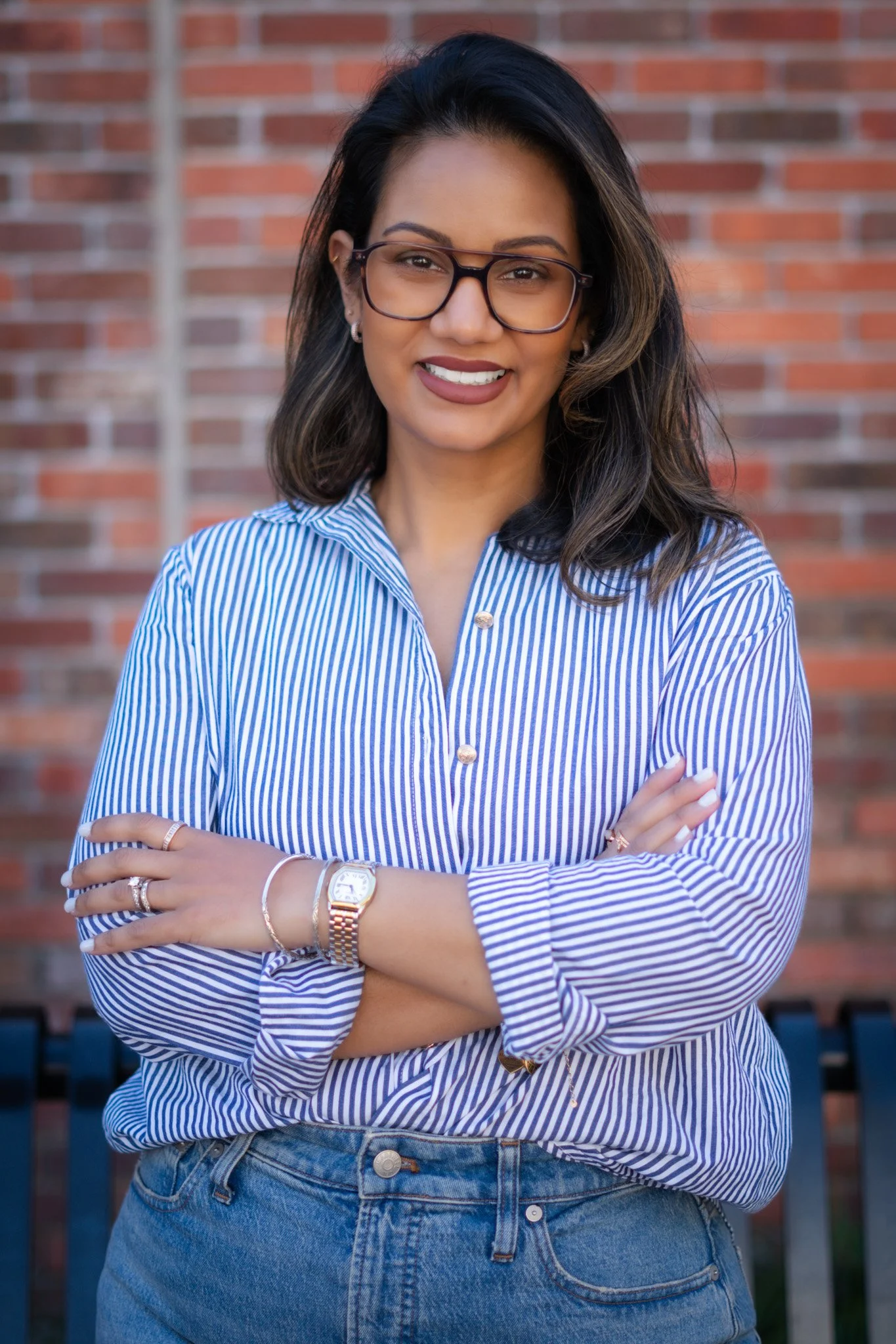 A woman with long dark hair wearing glasses and a blue and white striped shirt standing with arms crossed in front of a brick wall.