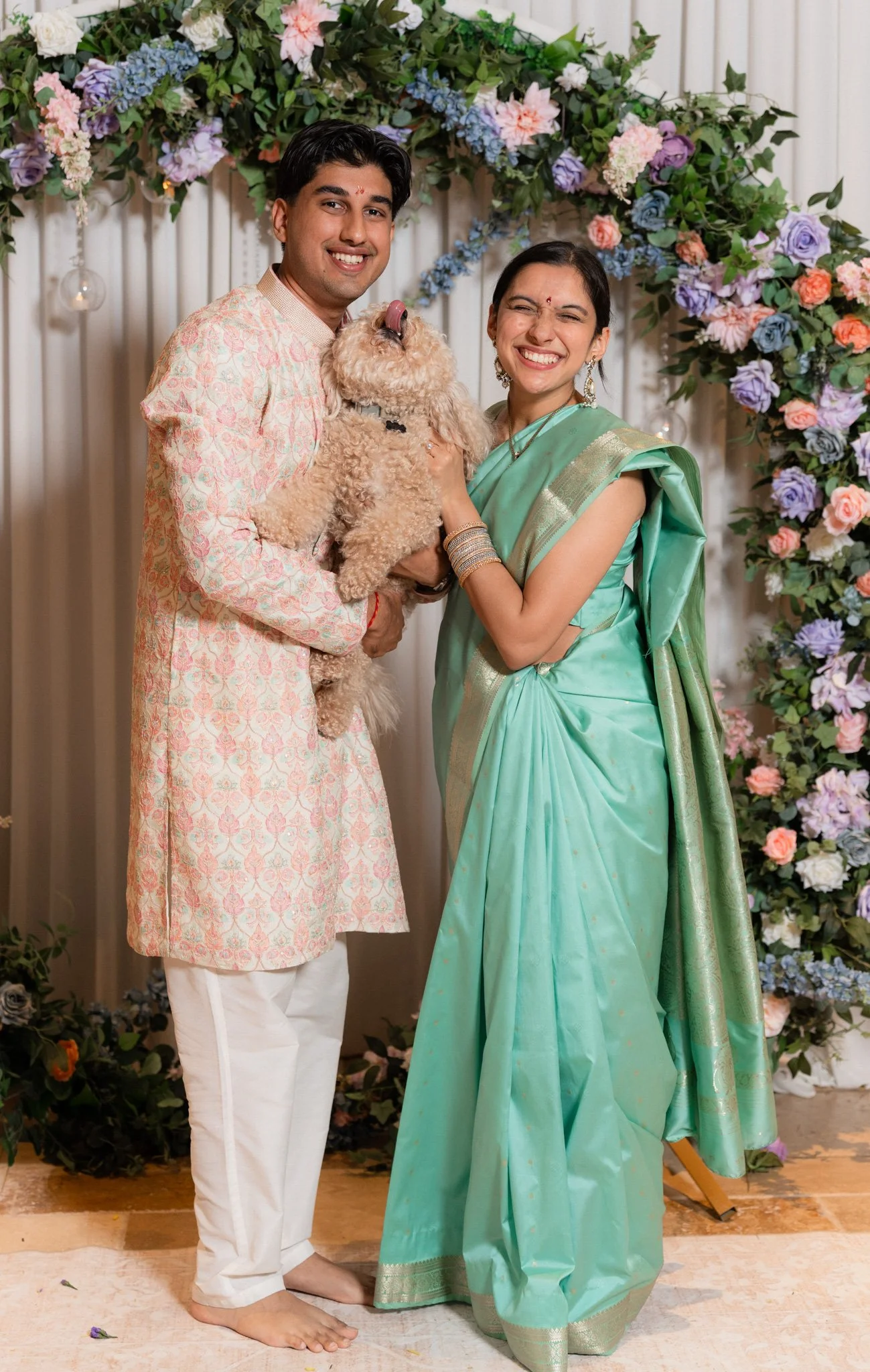 A man and woman smiling while holding a curly-haired dog in front of a floral arch at an indoor celebration. The man is wearing a pale pink and cream traditional outfit and white pants, and the woman is dressed in a green sari with gold accents. The background features draped curtains and colorful flowers.