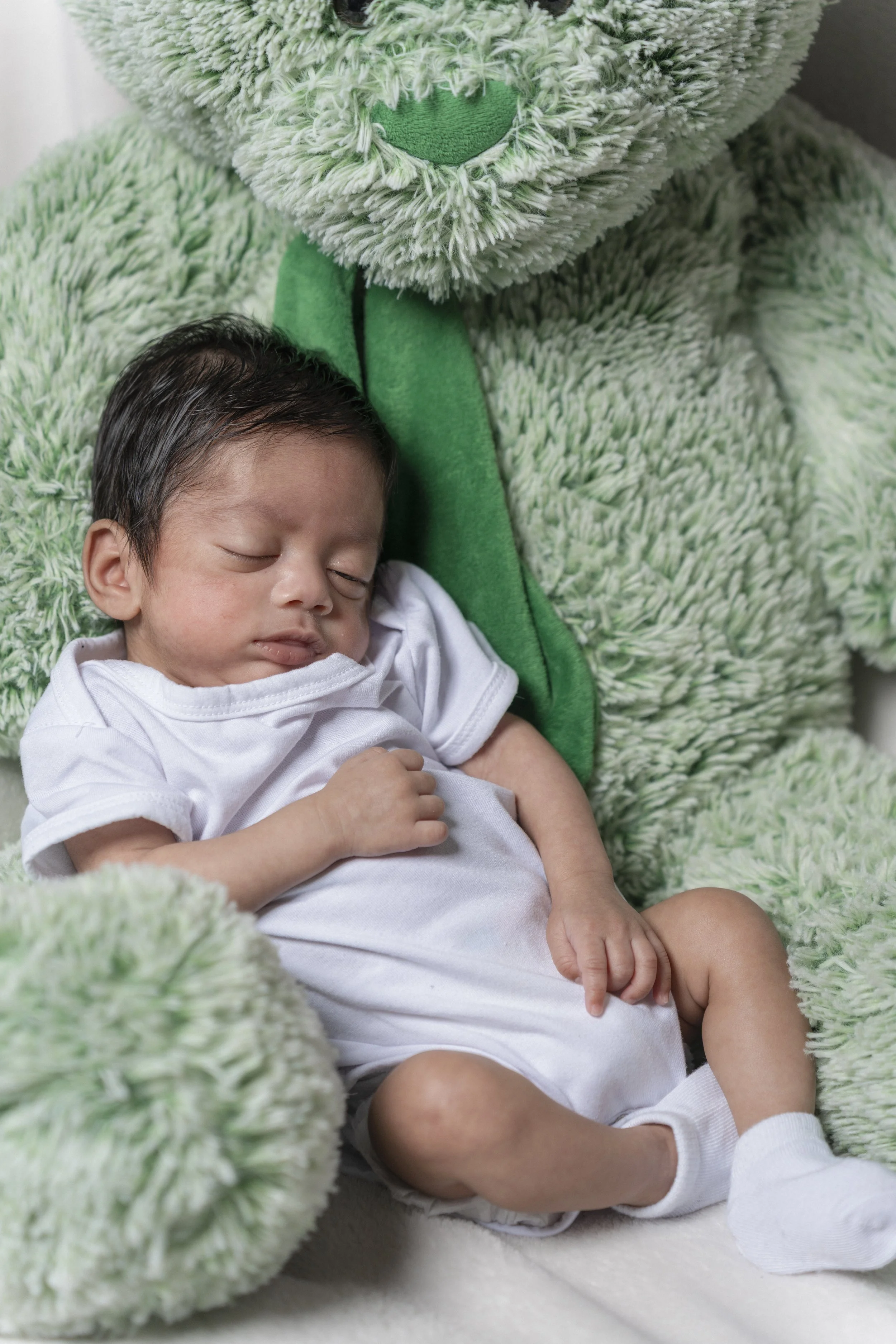 A sleeping baby lying on a soft surface, surrounded by a large, fuzzy, green plush teddy bear.
