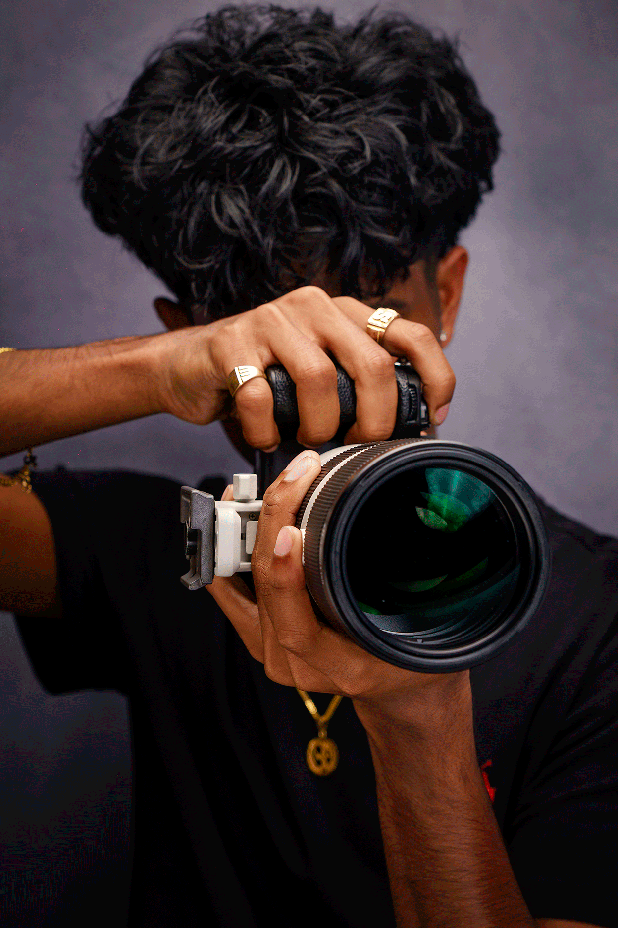 Person taking a photo with a professional camera, partially visible face, wearing rings and a gold necklace with a pendant, against a dark background.