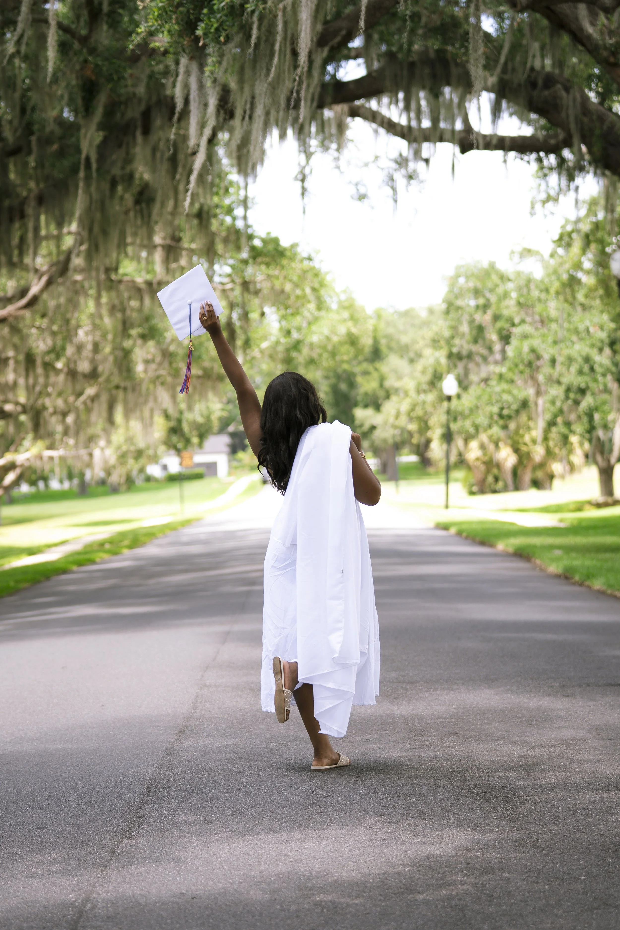 A young woman in a white dress and heels, celebrating her graduation, is standing on a paved road with her back to the camera, holding her diploma high in her right hand. She is outdoors in a park with green trees and hanging moss in the background.
