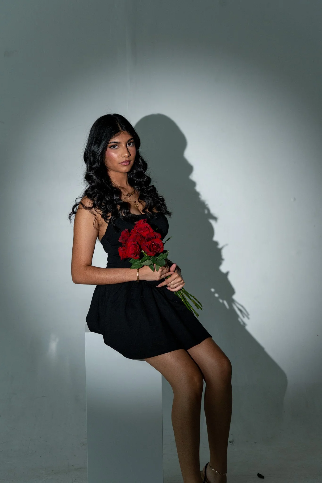 A woman with long wavy black hair, wearing a black dress, holding a bouquet of red roses, standing in front of a plain wall with a shadow cast to her right.