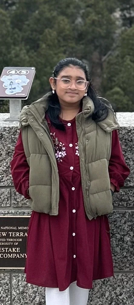 Young woman with glasses and earrings wearing a red dress, green puffer vest, and white leggings, standing outdoors with trees and a memorial plaque behind her.