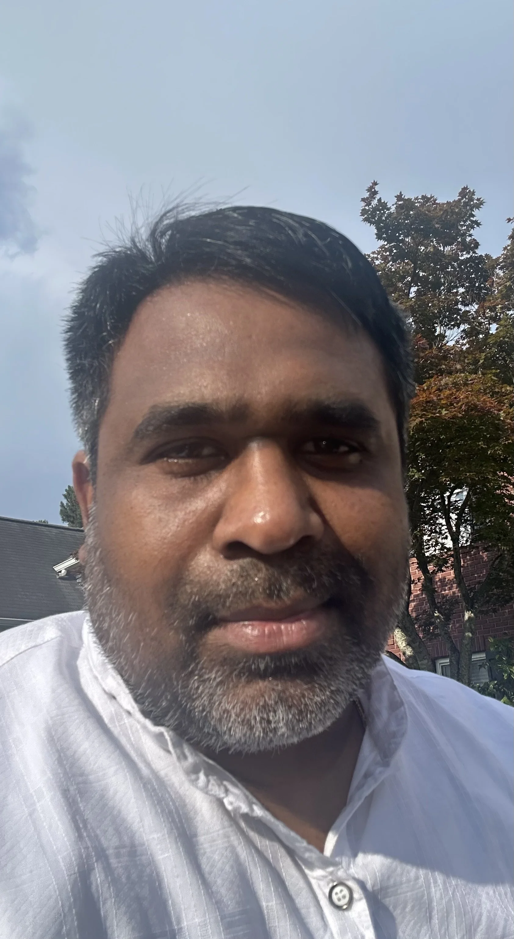 A close-up portrait of a man with medium skin tone, black and gray hair, and a beard, smiling outdoors in front of trees and a blue sky.