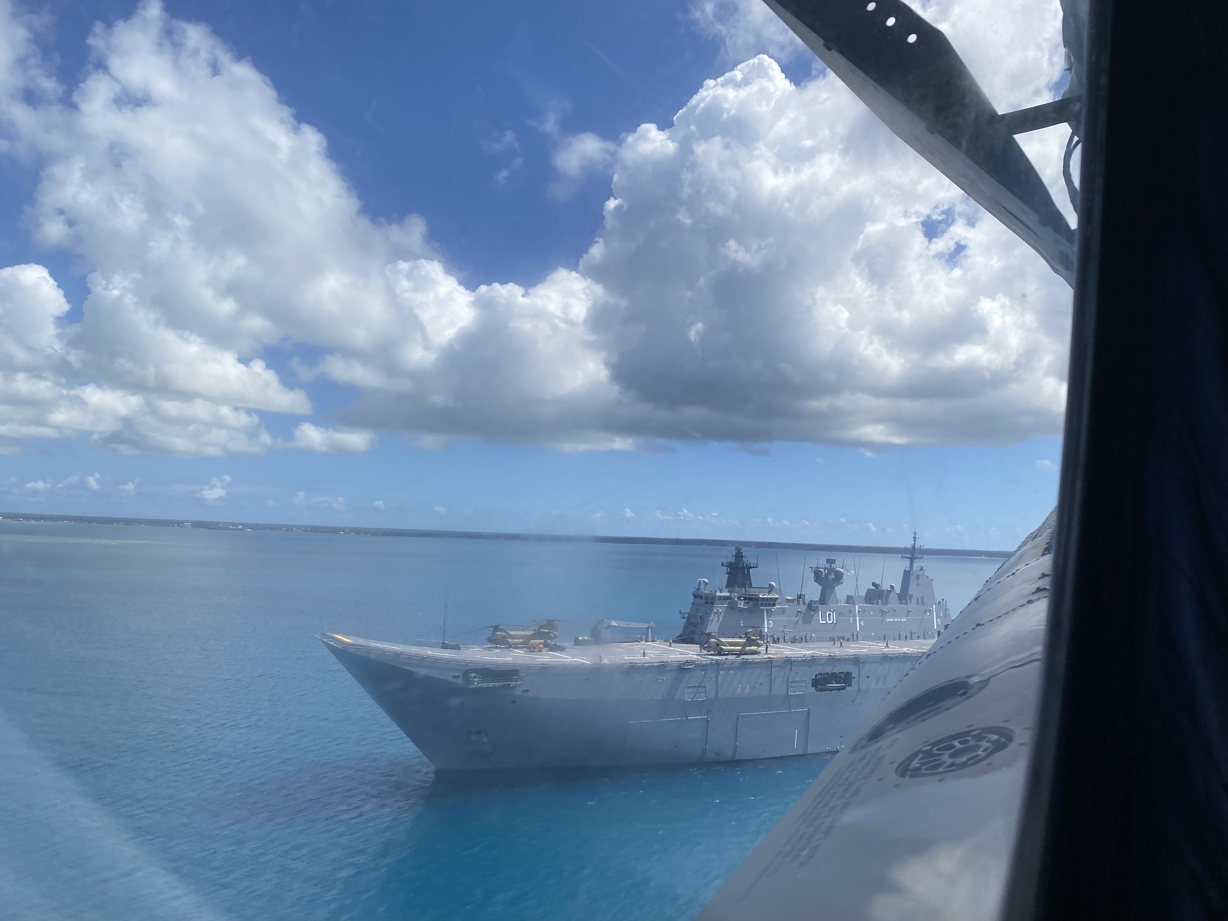 A large military ship docked at a port with a helicopter on the deck, under a partly cloudy sky.