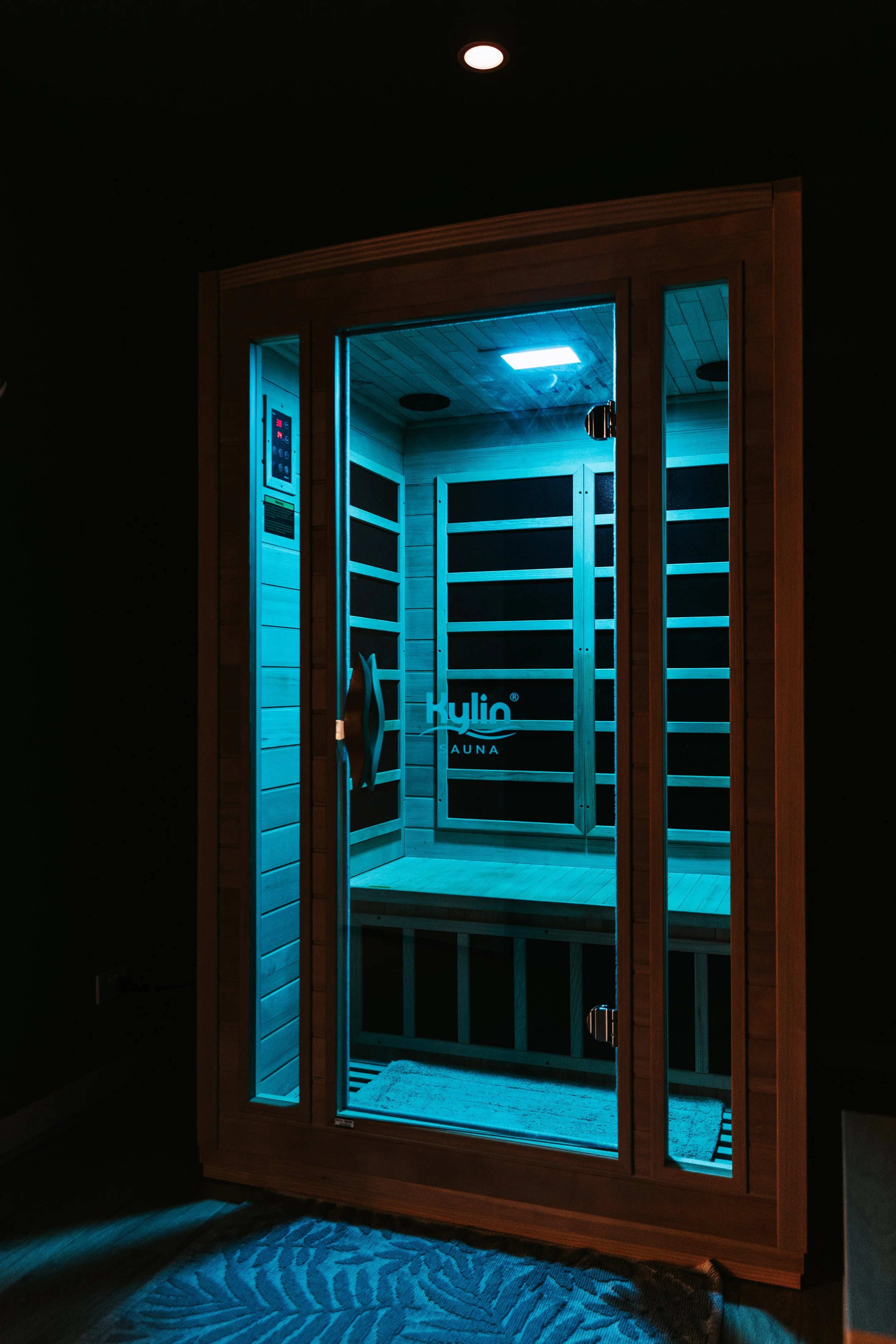 Sage Pilates Brunswick West © | A wooden sauna room illuminated with blue LED lighting, visible through a glass door with a wooden handle, and an information panel on the left wall.