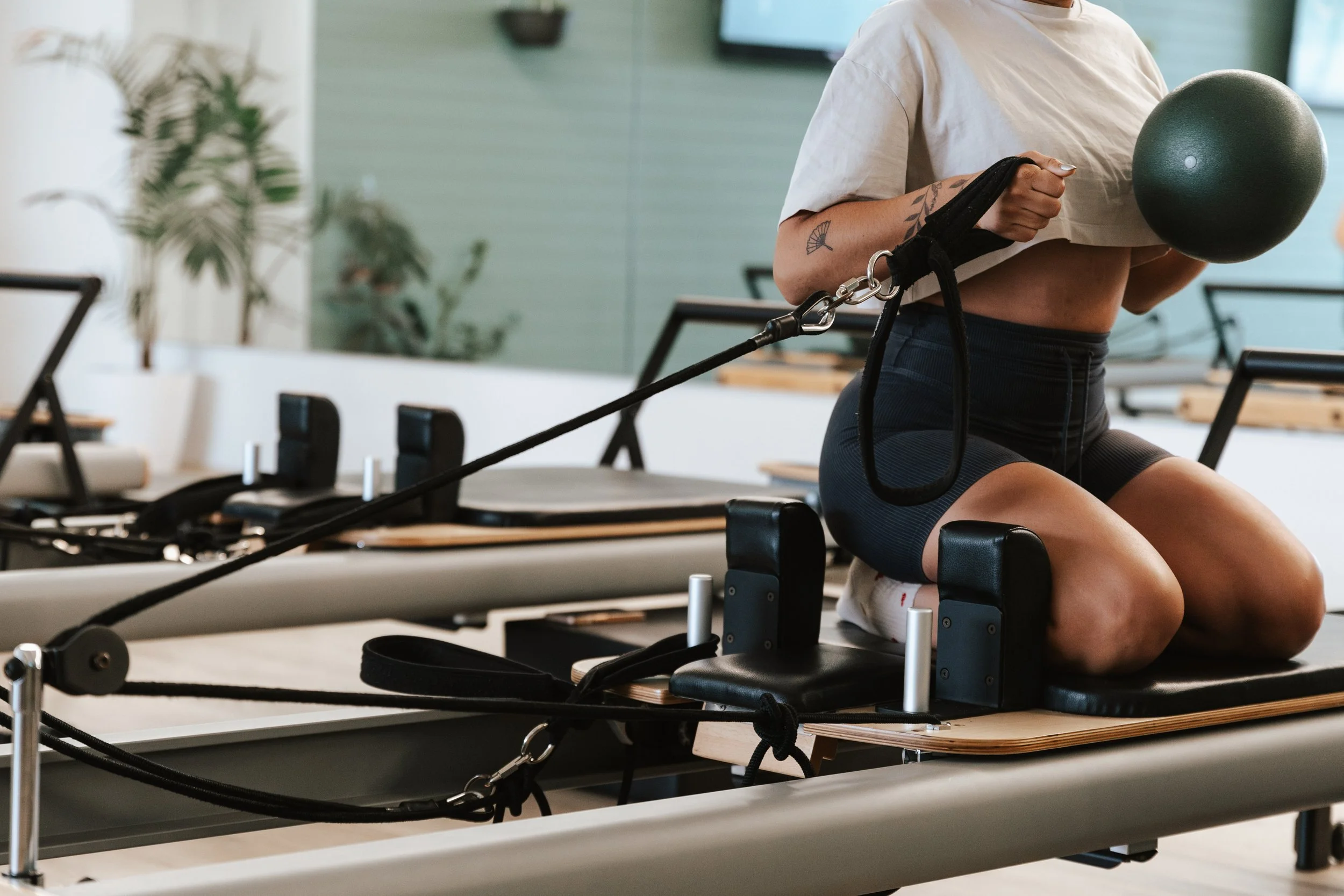 Sage Pilates Brunswick West © | A person sitting on a Pilates reformer machine in a fitness studio, holding a black exercise ball and wearing athletic clothing.