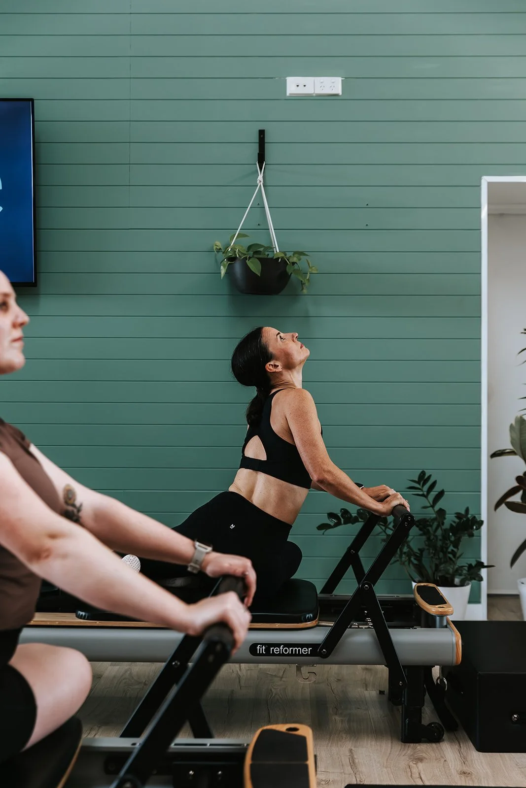 Sage Pilates Brunswick West © | People exercising on Pilates reformers in a fitness studio with a green wall and plants.