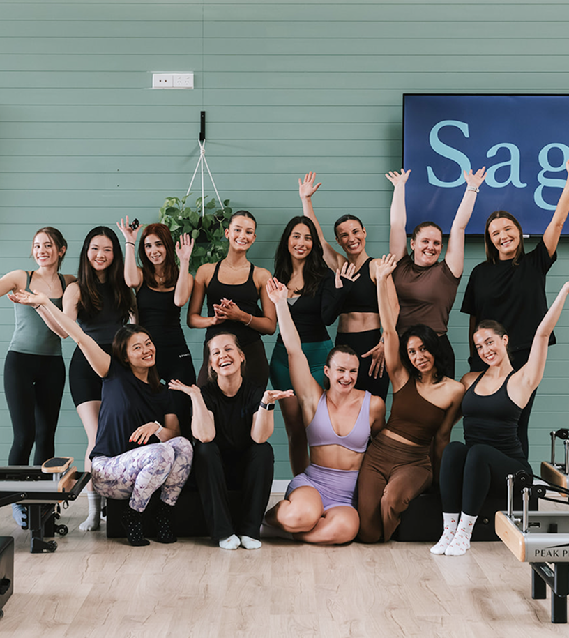 Sage Pilates Brunswick West © | A group of women in workout clothes posing and smiling in a fitness studio, some with arms raised, with a large digital screen displaying the word 'Sage' in the background.