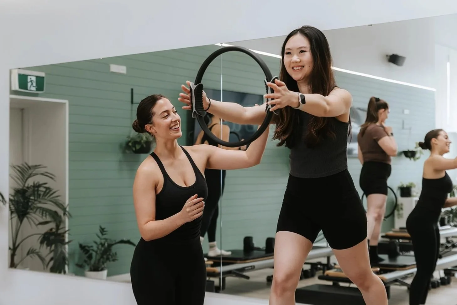 Sage Pilates Brunswick West © | Two women in workout clothes exchanging a fitness ring in a gym, with other women exercising in the background.