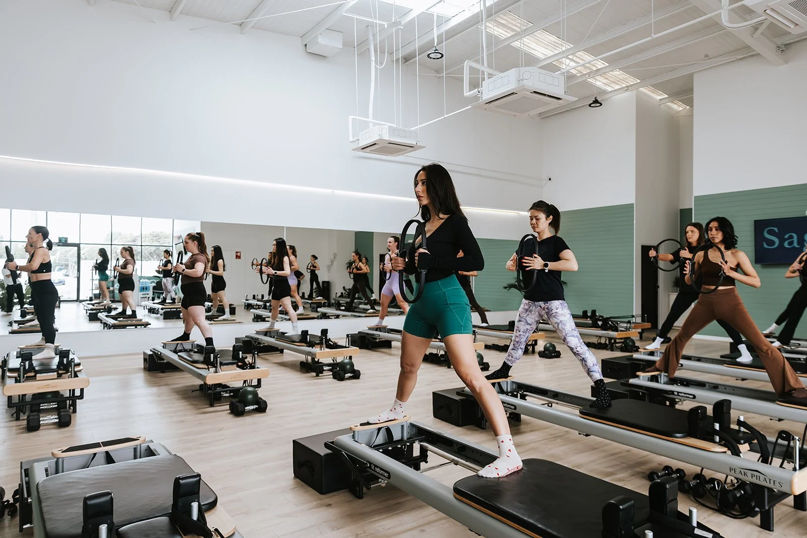 Sage Pilates Brunswick West © | A group of women participating in a Pilates class in a bright, spacious studio with large mirrors and wood floors, using reformer machines.