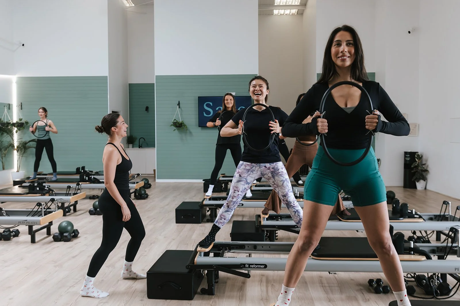 Sage Pilates Brunswick West © | A group of women participating in a fitness class on reformer Pilates machines in a bright, modern studio with light green walls and wooden floors. Some women are smiling and holding Pilates rings.
