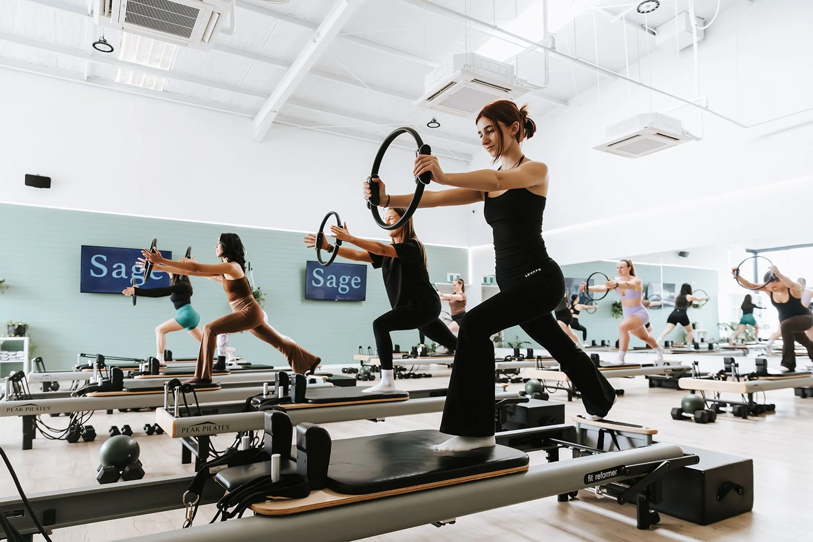 Sage Pilates Brunswick West © | A group of women participating in a Pilates class using reformer machines in a bright, spacious studio. They are all holding Pilates rings and appear focused on their exercise routines.