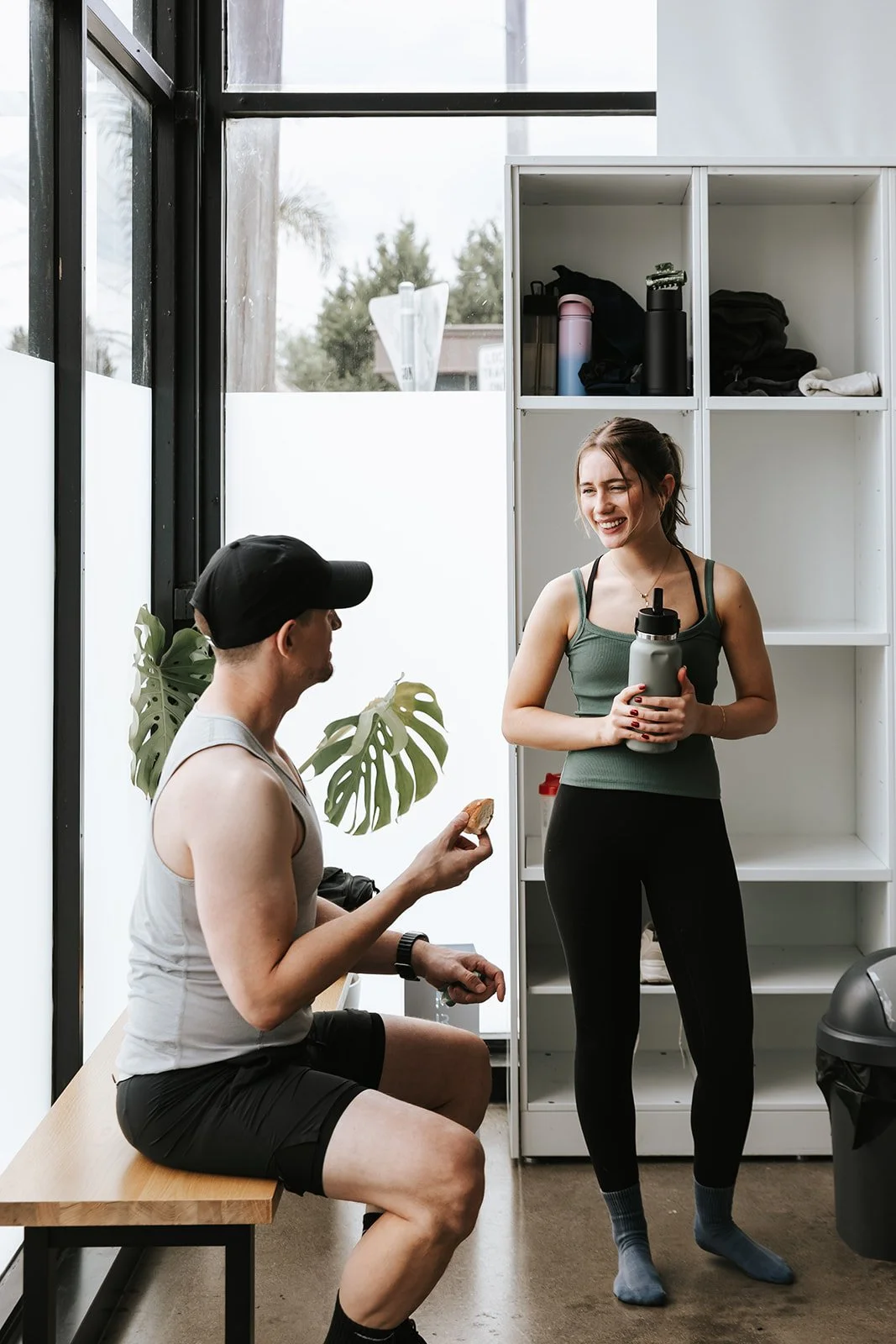Sage Pilates Brunswick West © | Two people conversing in a gym locker room, one seated on a bench holding a snack and wearing a black cap, the other standing with a water bottle, smiling.