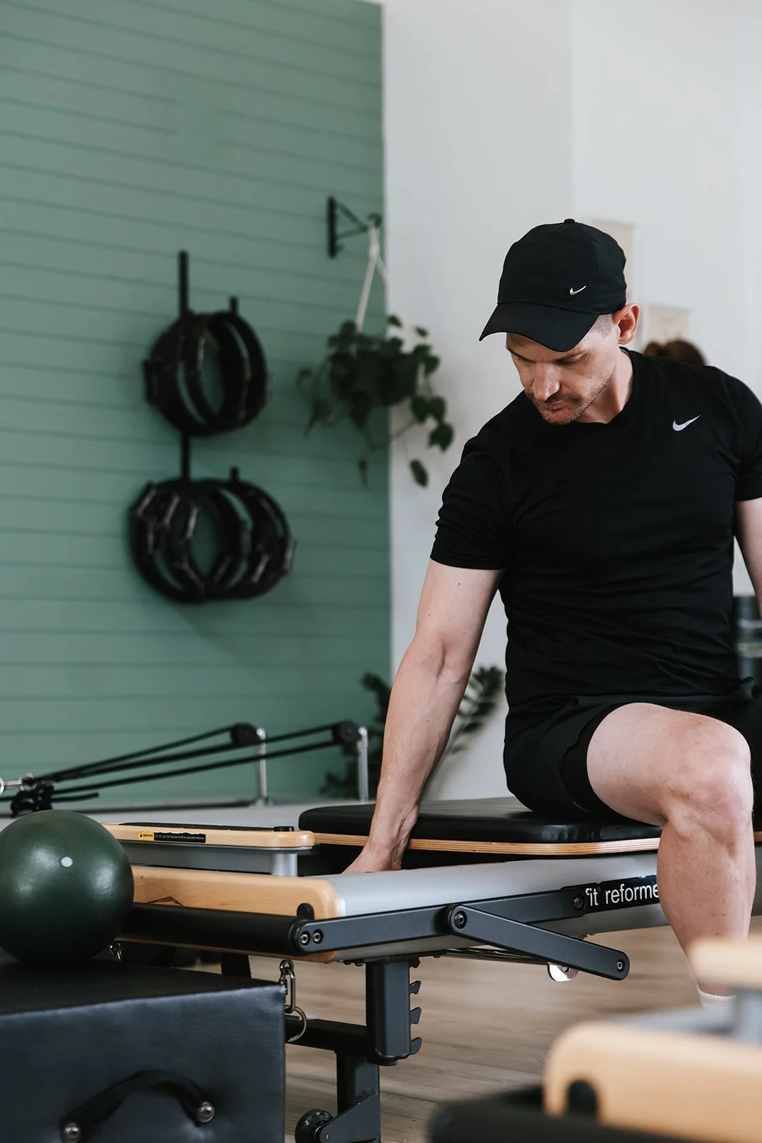 Sage Pilates Brunswick West © | A man exercising on a Pilates reformer machine in a fitness studio. He is wearing a black Nike cap and black athletic clothing.