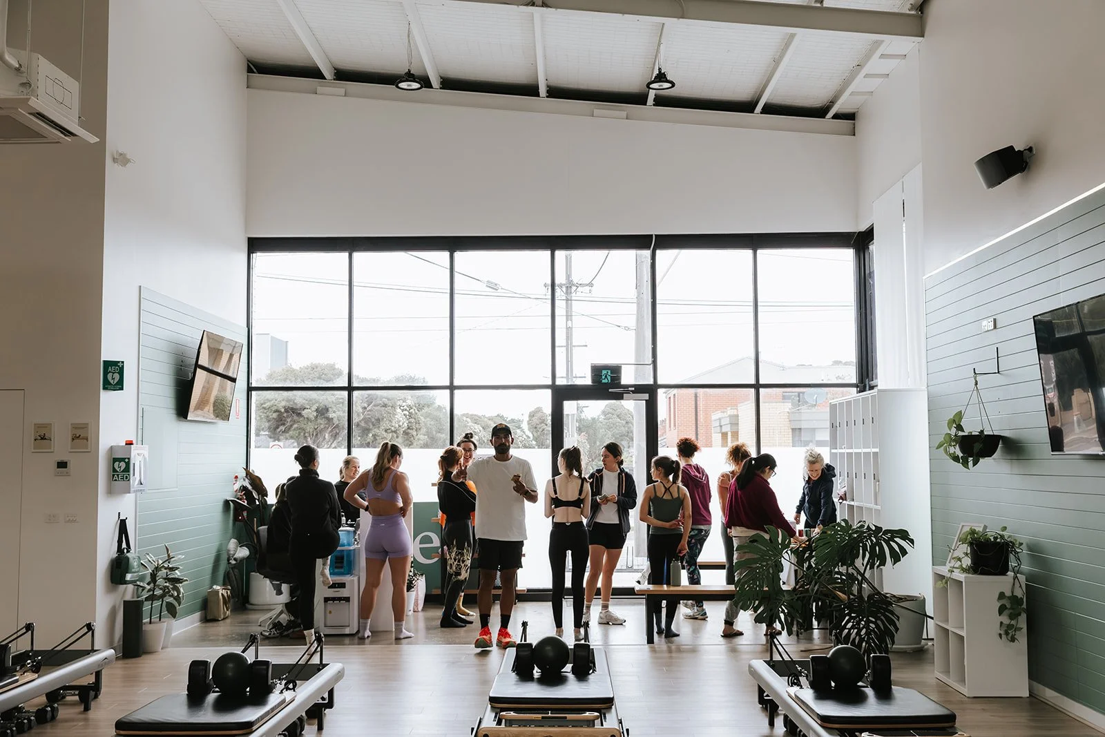Sage Pilates Brunswick West © | A group of people in workout attire standing in a fitness studio with large windows, some talking and others looking at their phones. The studio has exercise equipment, plants, and a wall-mounted TV.