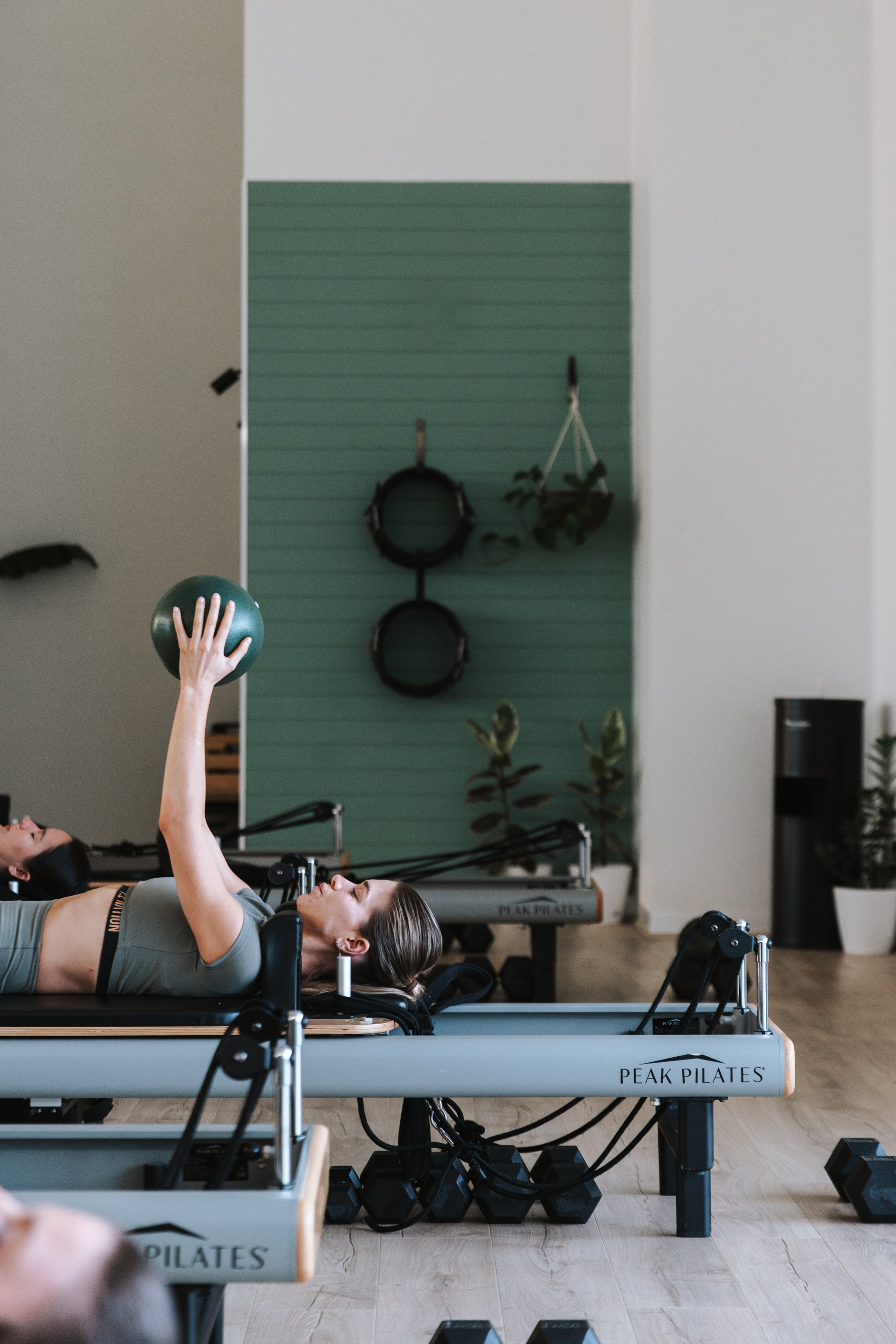 Sage Pilates Brunswick West © | Two women lying on Pilates reformer machines in a fitness studio, lifting small exercise balls above their heads, with a green wall and hanging fitness equipment in the background.