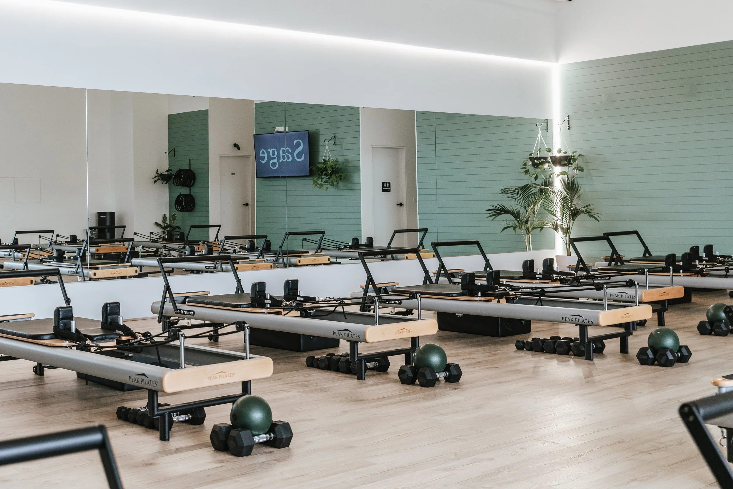Sage Pilates Brunswick West © | Pilates studio with reformer machines, dumbbells, and green exercise balls. The room has a wooden floor, a large mirror on the wall, and plants for decor.