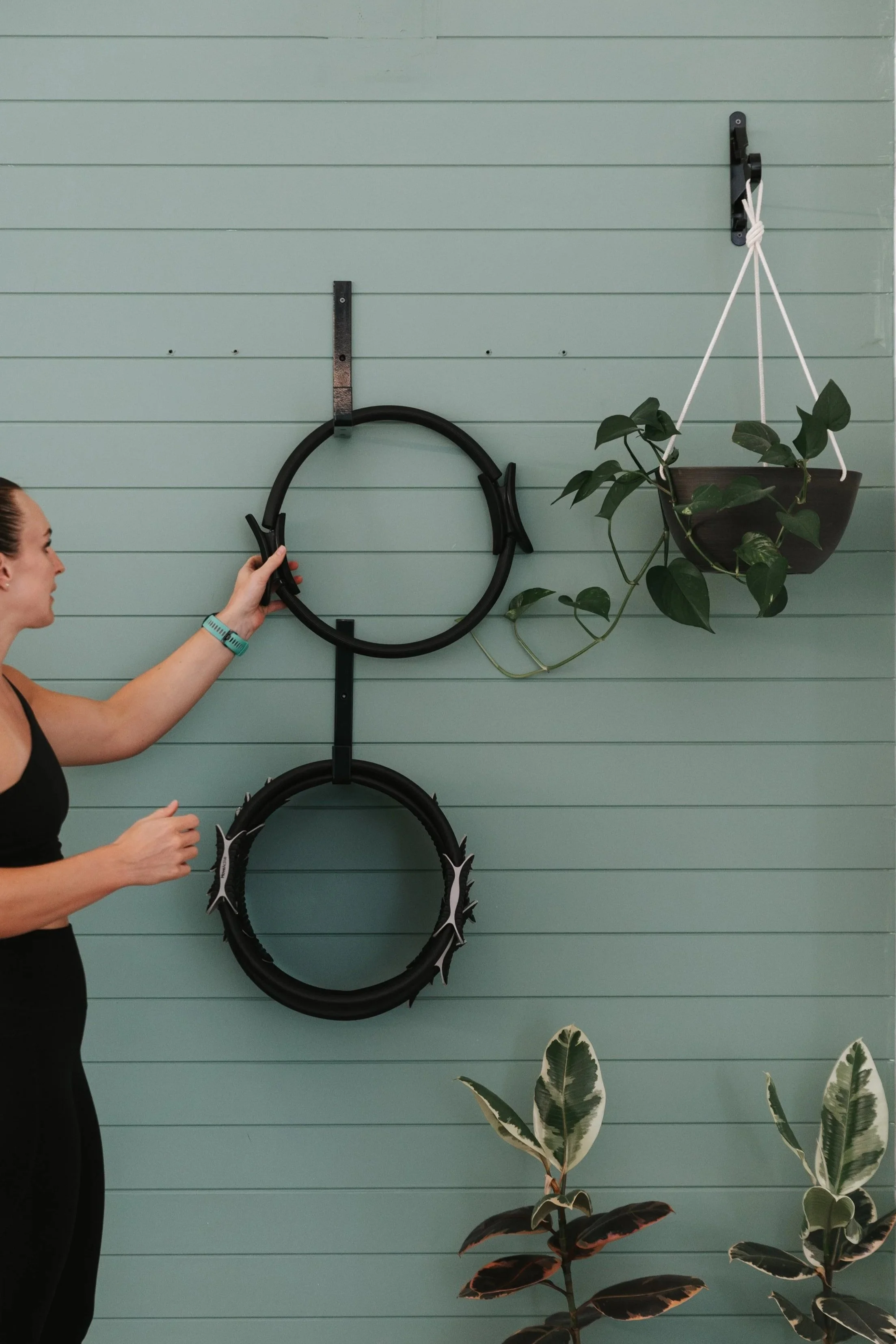 Sage Pilates Brunswick West © | Woman attaching a black bicycle wheel to a blue wall with horizontal panels, next to a hanging potted plant with large green leaves, and other indoor plants below.