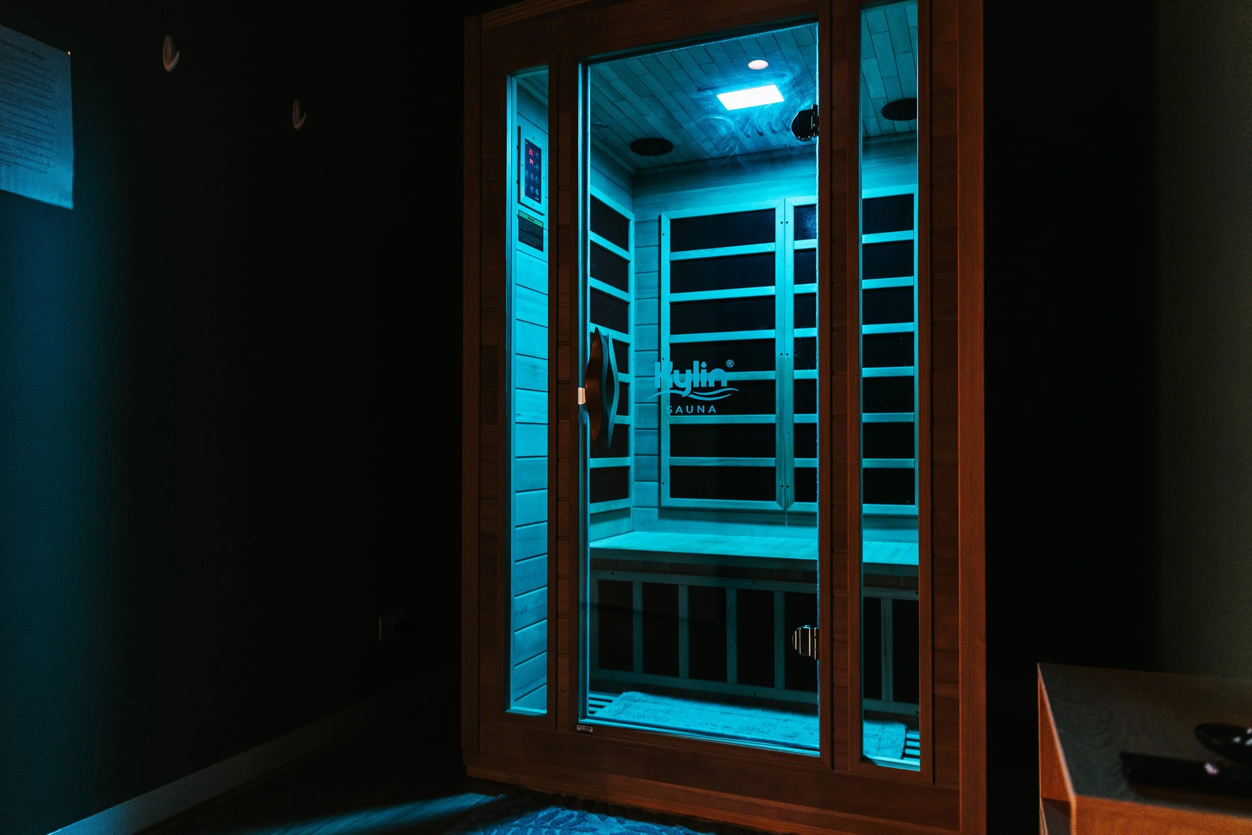 Sage Pilates Brunswick West © | A wooden sauna room with closed glass door, illuminated with blue light, showing wooden interior and black heating panels.