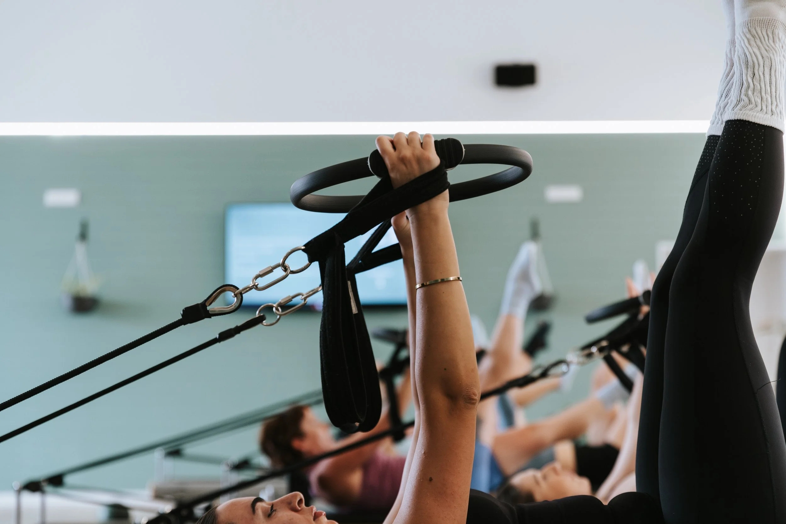 Sage Pilates Brunswick West © | Participants in a fitness class are lying on mats and using suspension training equipment, with arms raised holding handles. The scene is in a bright, modern gym or fitness studio.