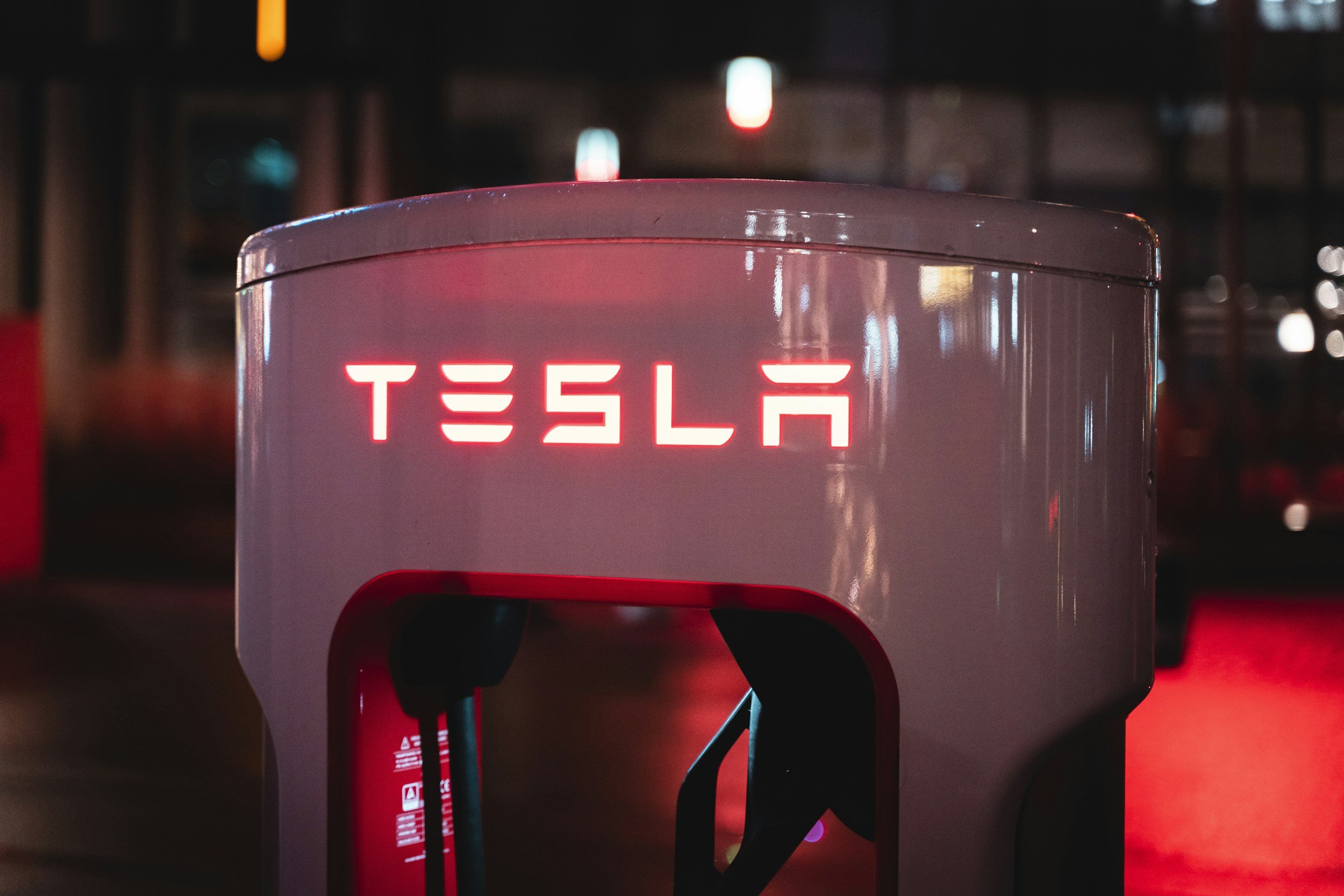 A Tesla electric vehicle charging station illuminated at night with a red glow, showing the partially visible Tesla logo, surrounded by darkness and some blurred lights in the background.