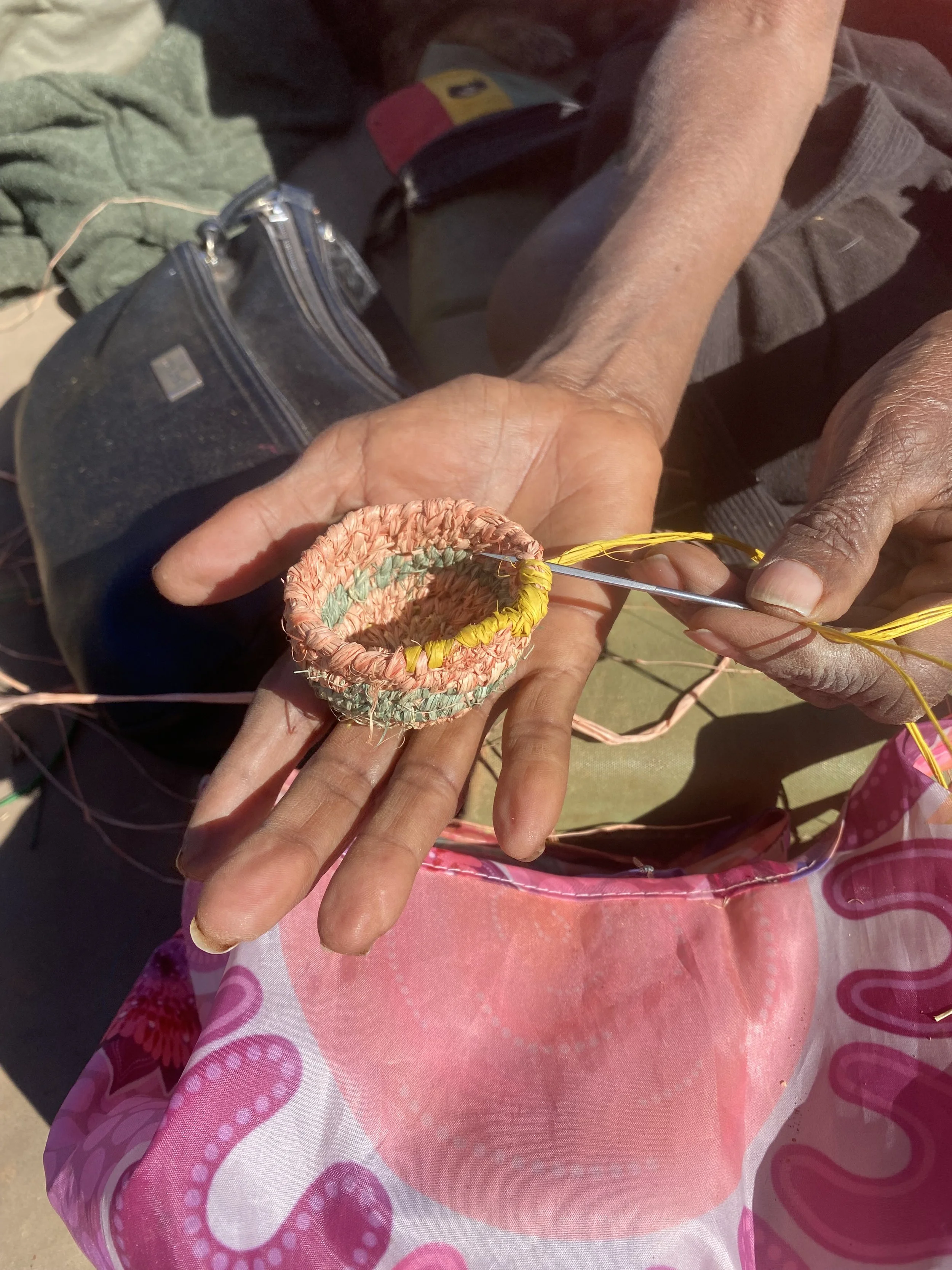 Close-up of hands weaving a small basket with colorful materials, with a child sitting nearby wearing pink and purple clothing.