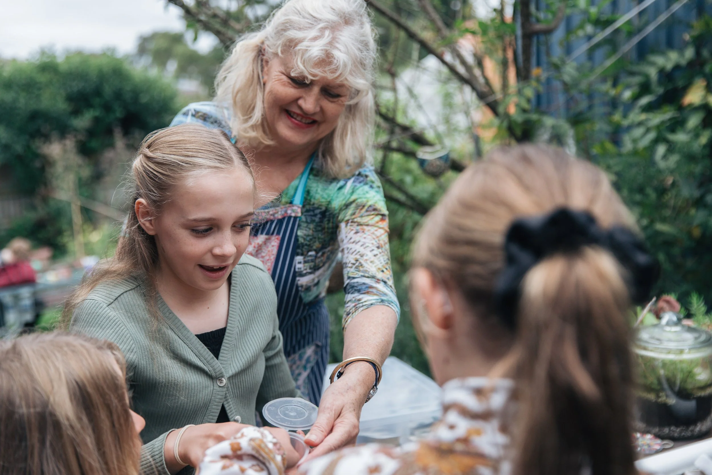 A woman and a girl enjoying a smile while sitting outside at a table, surrounded by other children, during daytime with green trees in the background.
