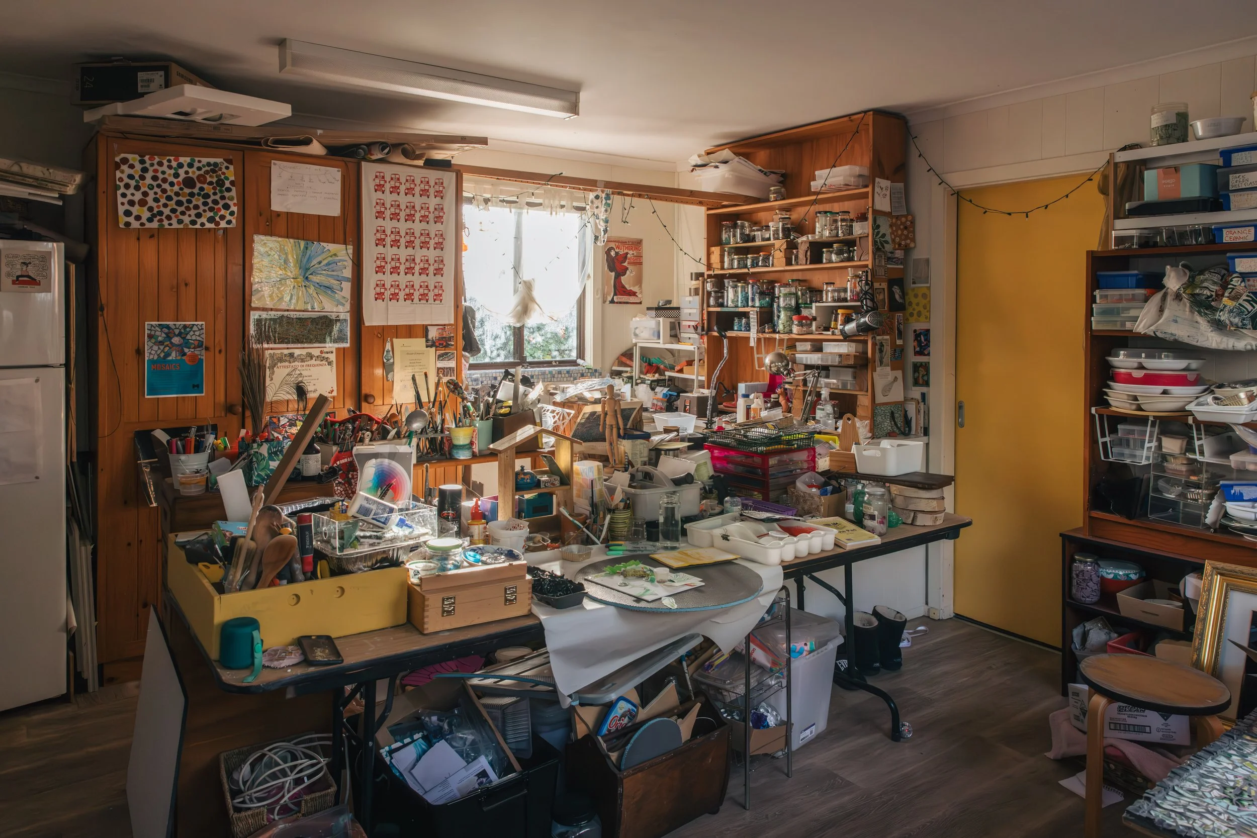 A cluttered craft room or studio with tables and shelves filled with art supplies, jars, papers, and tools. A window with string lights and a yellow door are visible in the background.