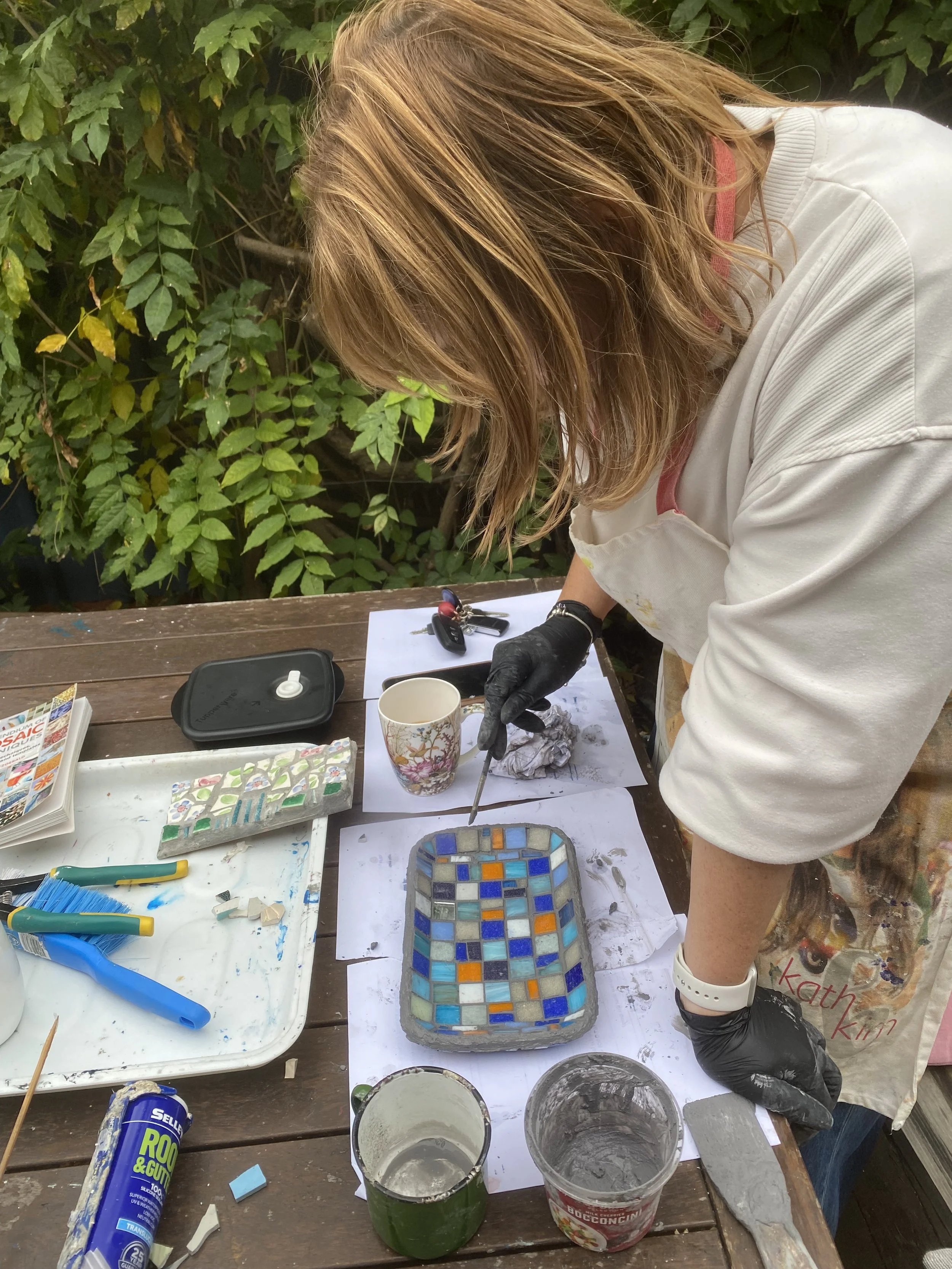 Person working on creating a mosaic artwork outdoors, with tools, containers of grout or adhesive, and a mosaic tile piece on the table, surrounded by green foliage.