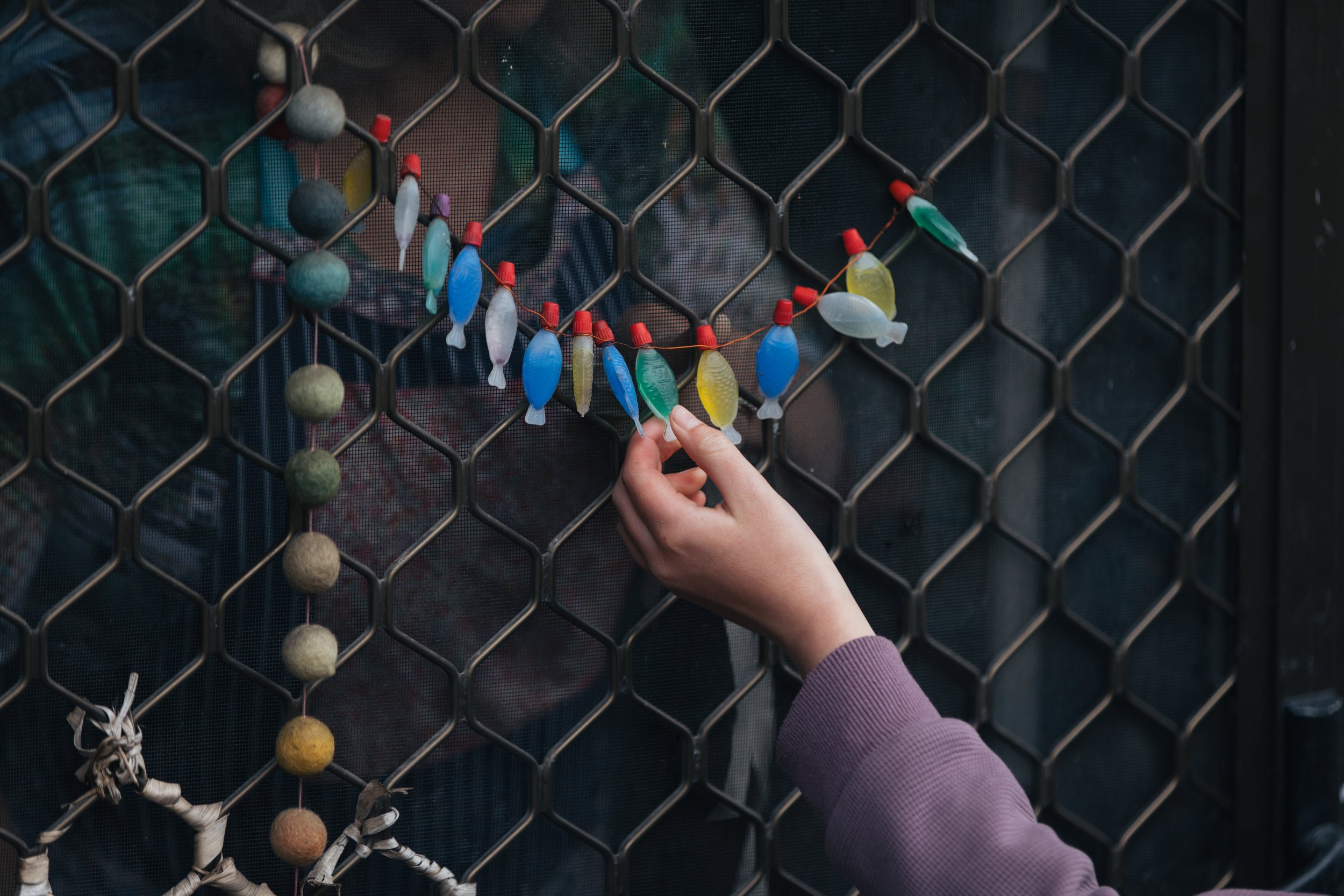 A person's hand adjusting a string of colorful fishing bobbers nailed to a wire fence.