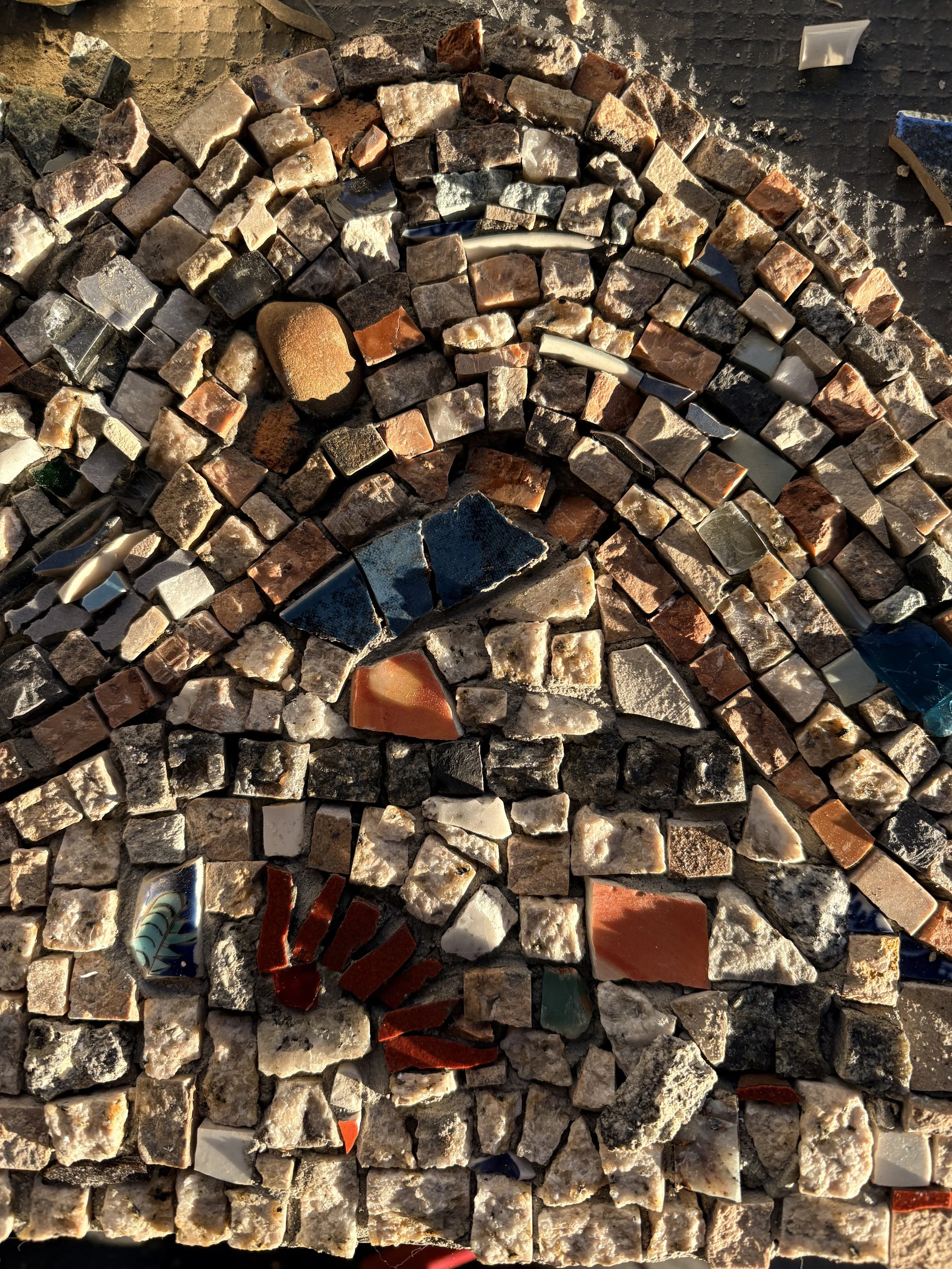 Close-up view of a mosaic wall composed of irregularly shaped stones and ceramic pieces in various colors such as white, beige, red, black, and blue. The stones are arranged to create a pattern with some small gaps filled with grout. Sunlight illumin
