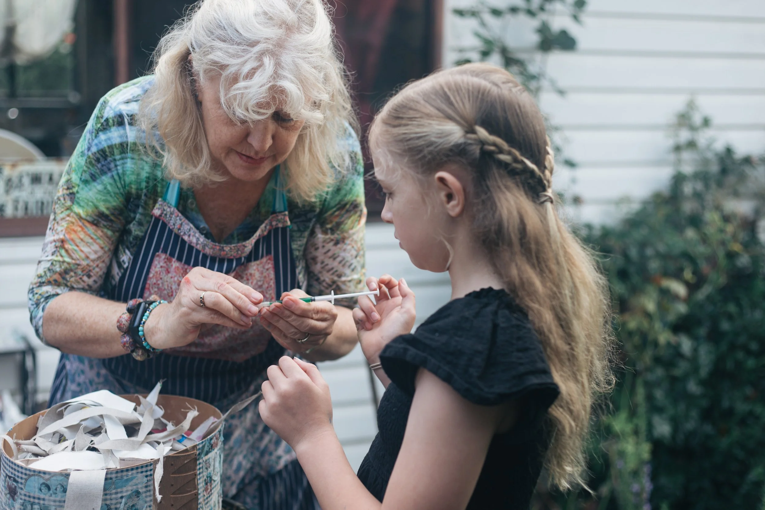 An elderly woman and a young girl are engaged in a craft activity outdoors, with the woman helping the girl with a craft project. The woman has white, curly hair and is wearing a colorful shirt and an apron, while the girl has long, light brown hair 