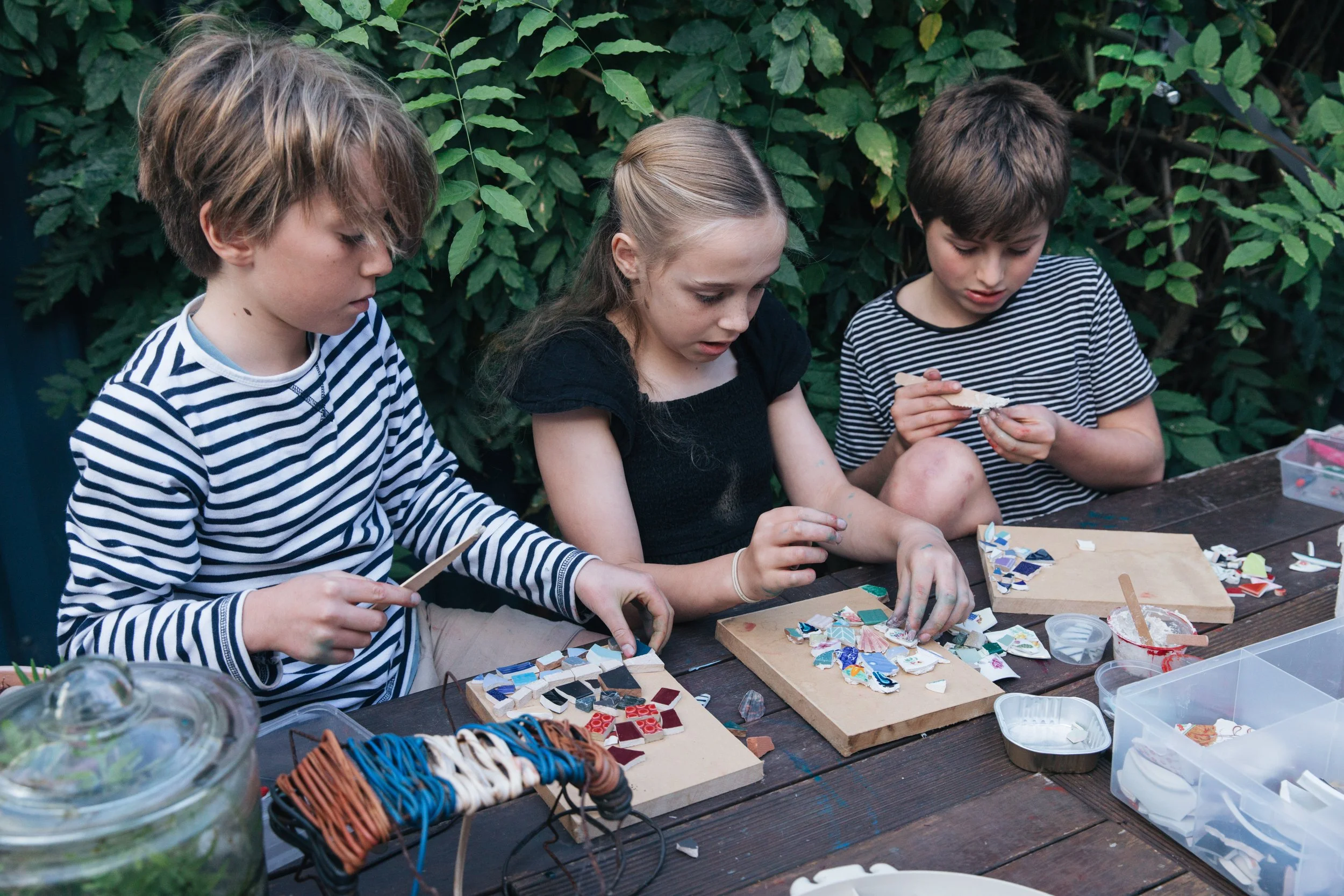 Three children sitting at a wooden table outdoors, working on mosaic art projects with various small colored tiles, amidst lush green foliage.