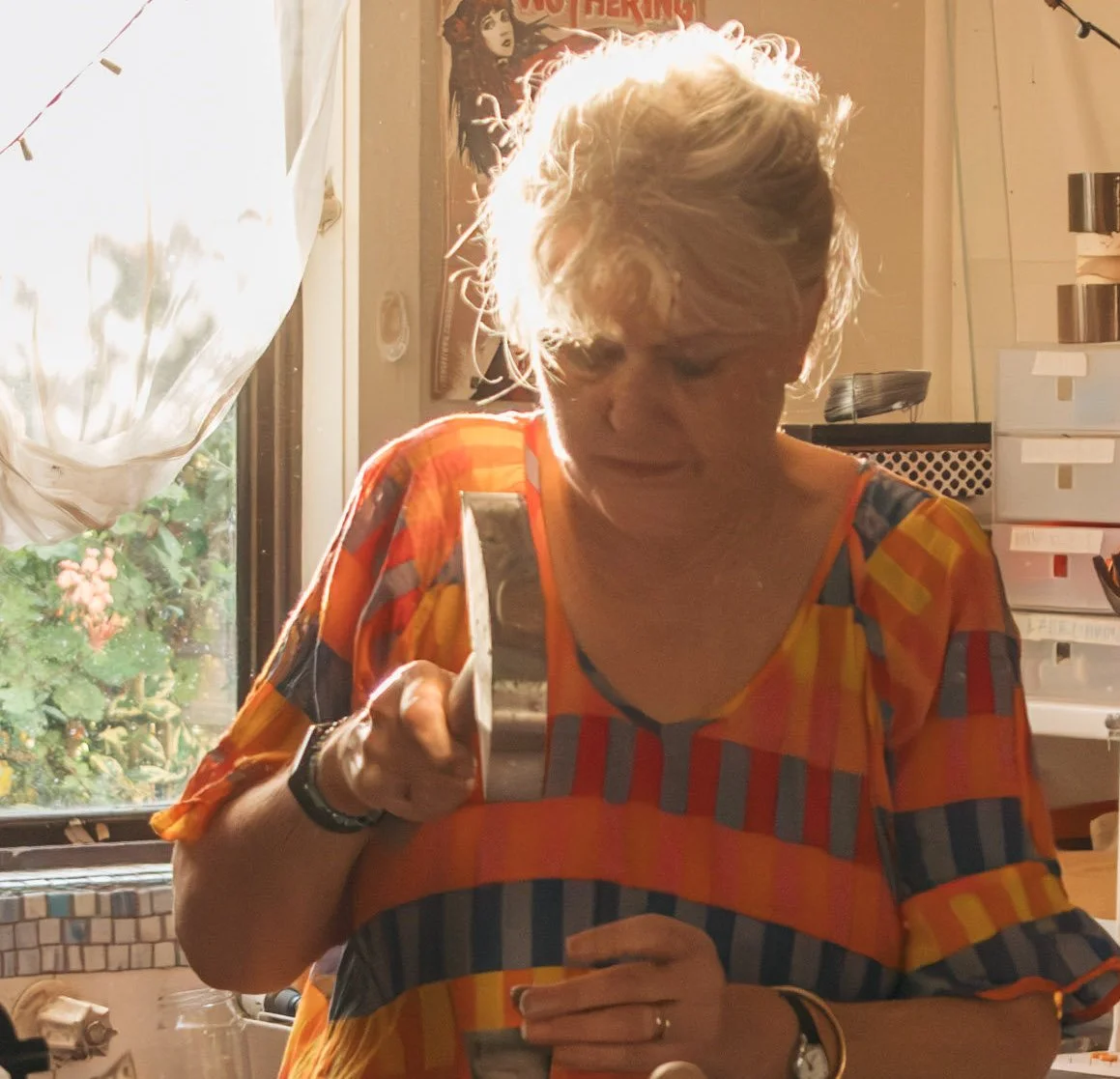 An older woman with short, curly, blonde hair is standing by a window with sunlight shining on her. She is wearing a colorful, striped shirt and holding a small object, perhaps a piece of food, in one hand while looking down at her phone in the other
