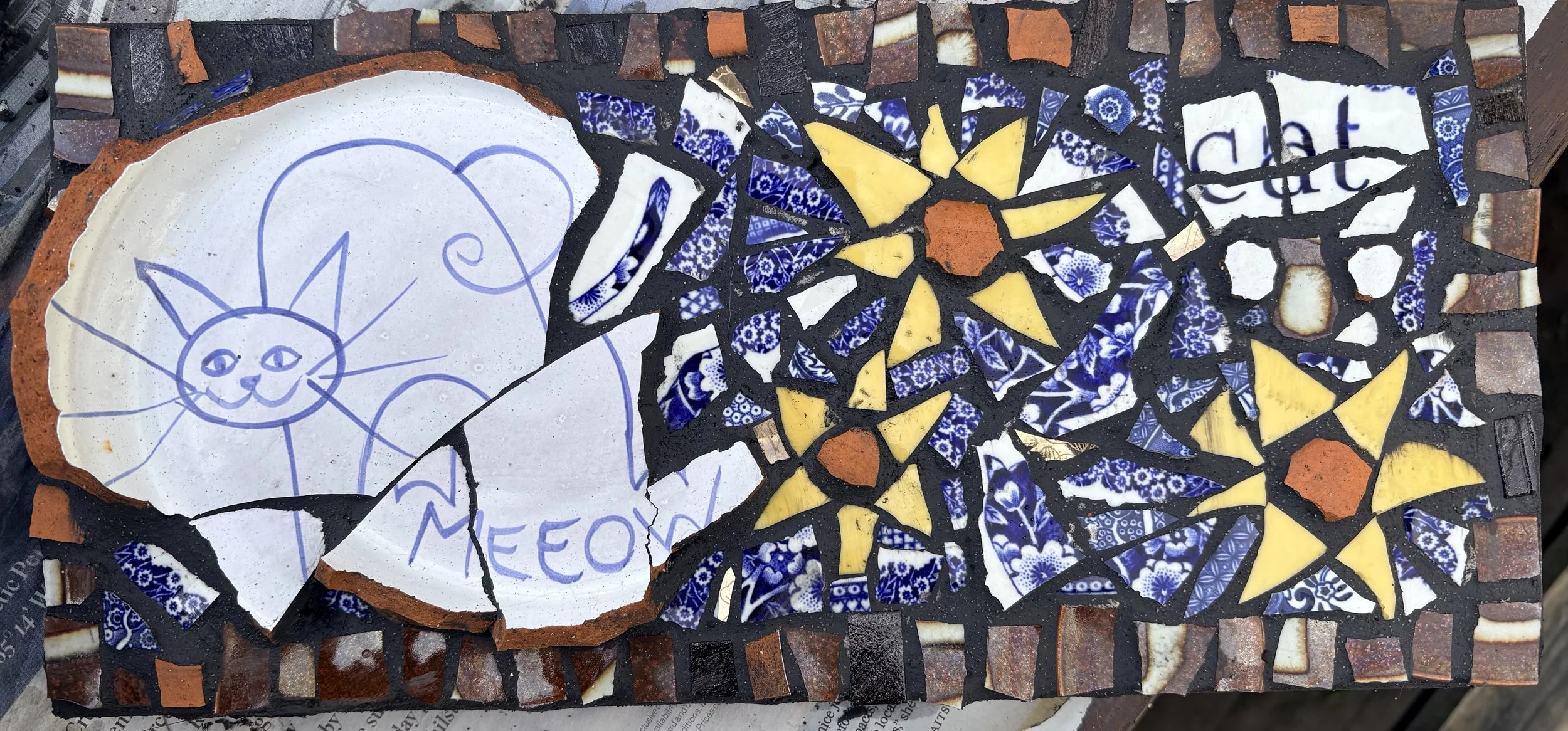 A mosaic made of broken ceramic pieces, featuring a drawing of a smiling cat with the word 'Meow' on a white tile, surrounded by blue and yellow decorative tiles.