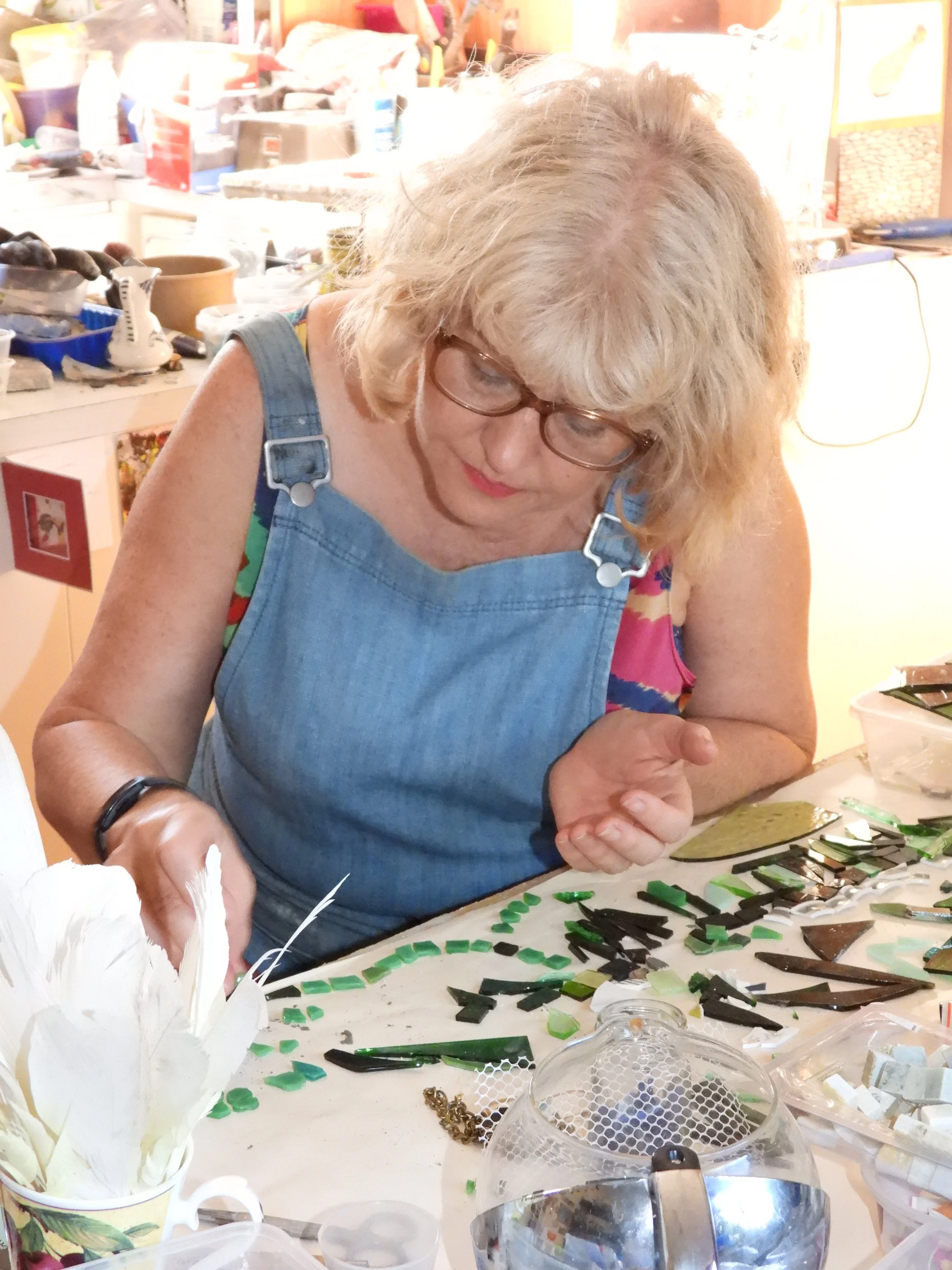 Woman working on a mosaic project with small pieces of green and black glass, surrounded by art supplies and tools, in a cluttered craft room.