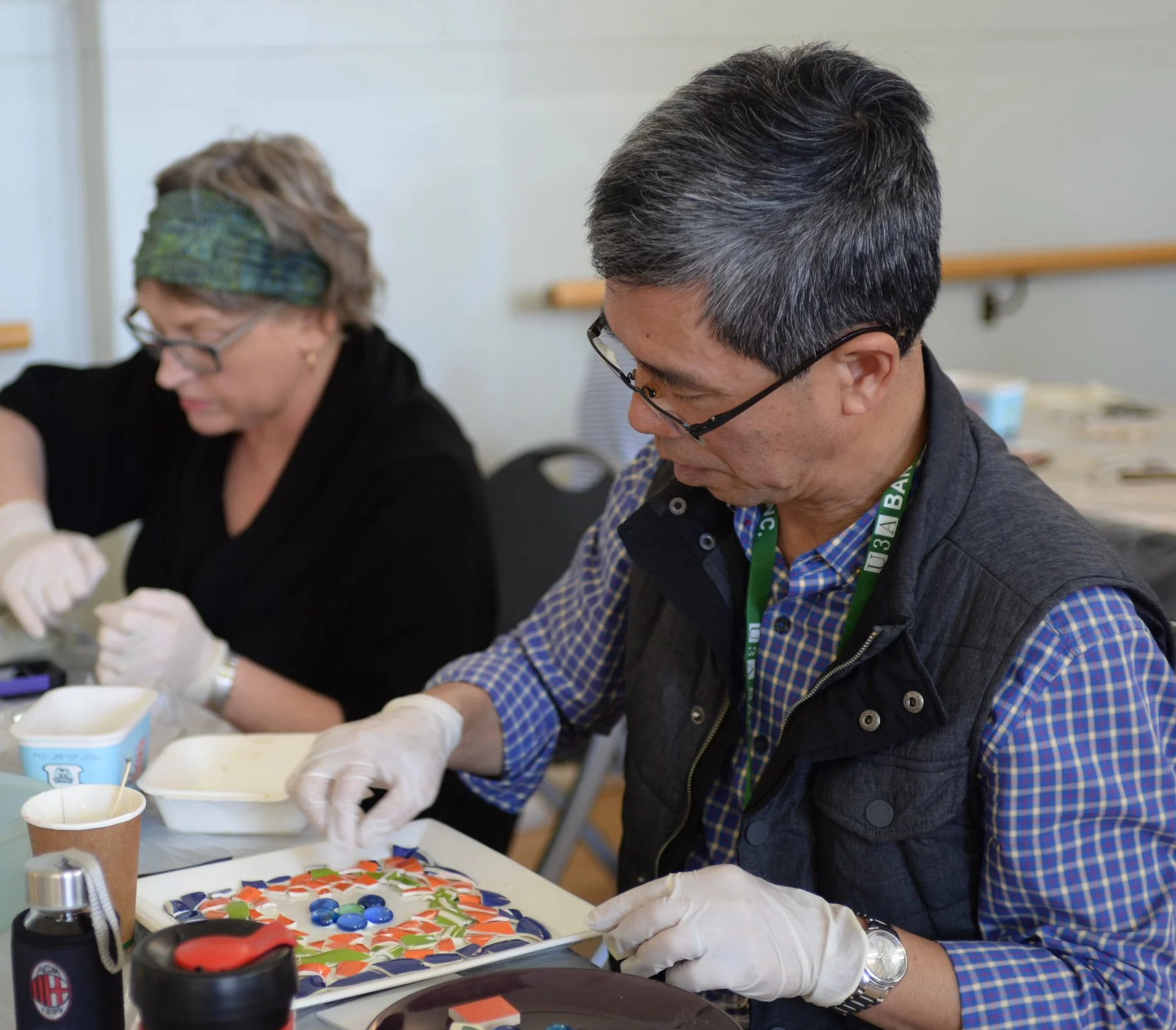 Two people working on a mosaic project at a table, wearing gloves and glasses, with colorful tiles and mosaic pieces in front of them in a room with a whiteboard in the background.