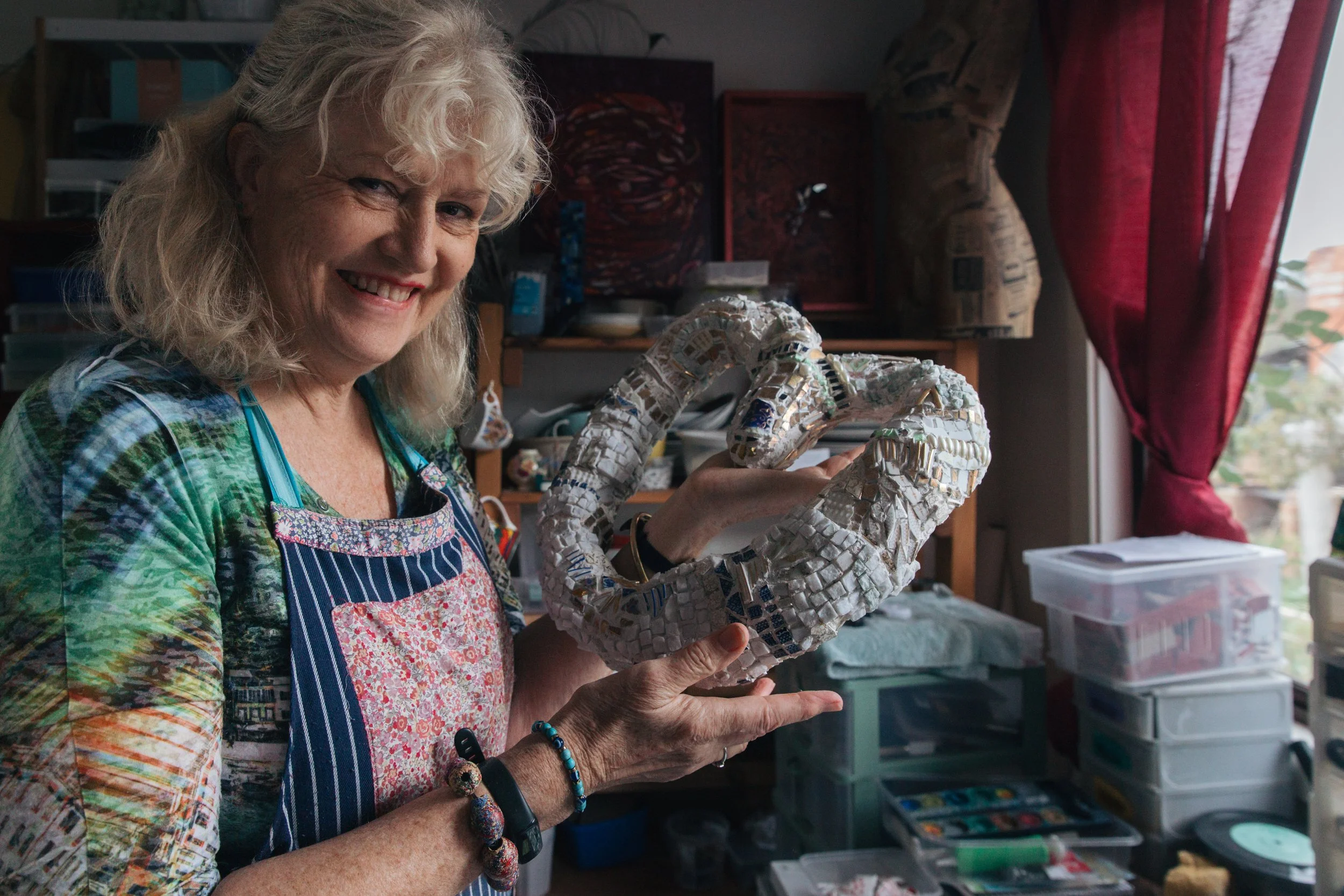 An elderly woman with curly gray hair is smiling and holding a large mosaic sculpture of a chameleon in her hands. She is wearing a colorful shirt and a striped apron, and is surrounded by art supplies and storage shelves in her art studio.