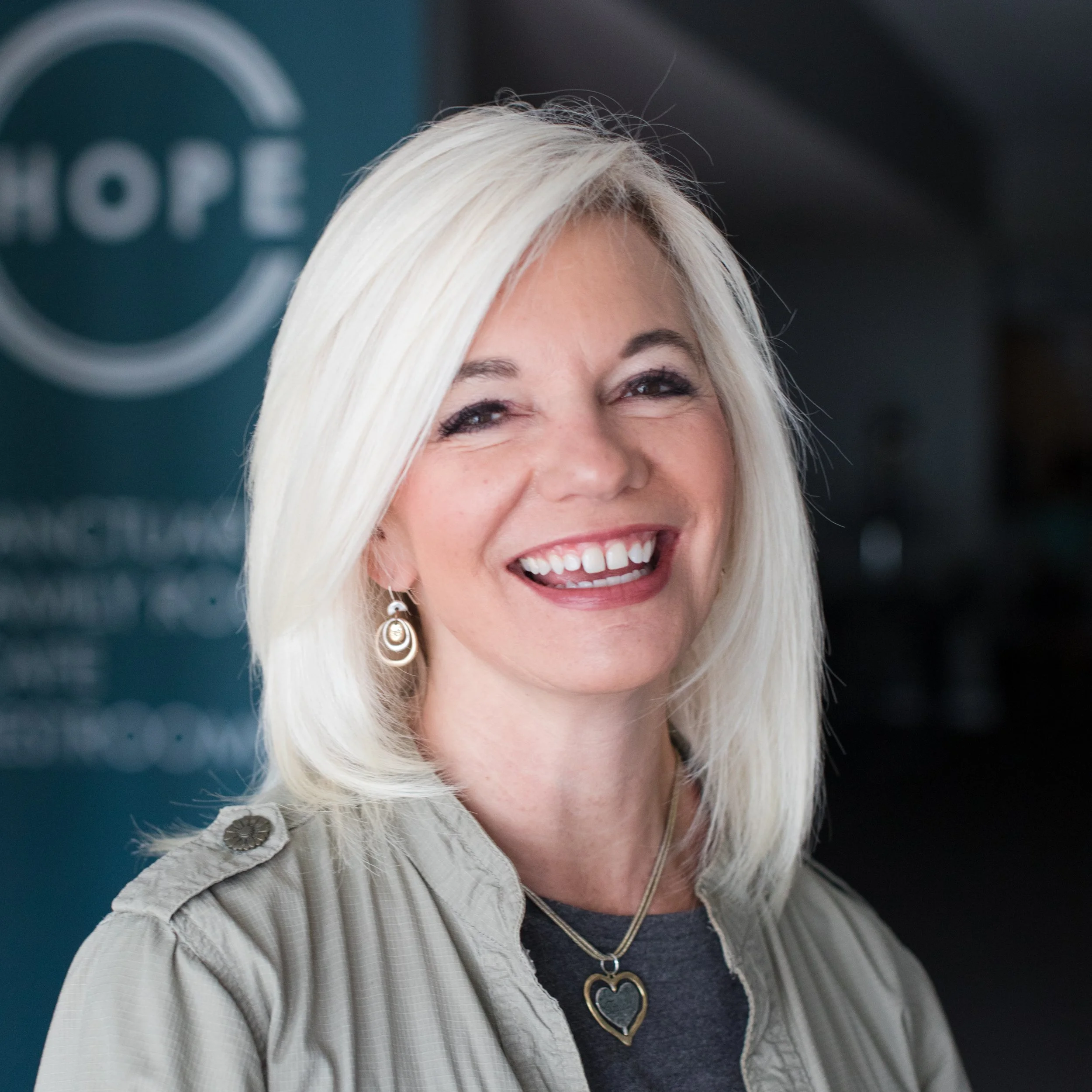 Close-up of a smiling woman with platinum blonde hair, wearing a gray shirt, layered necklaces, and earrings, with a blurred background showing the word 'HOPE'.