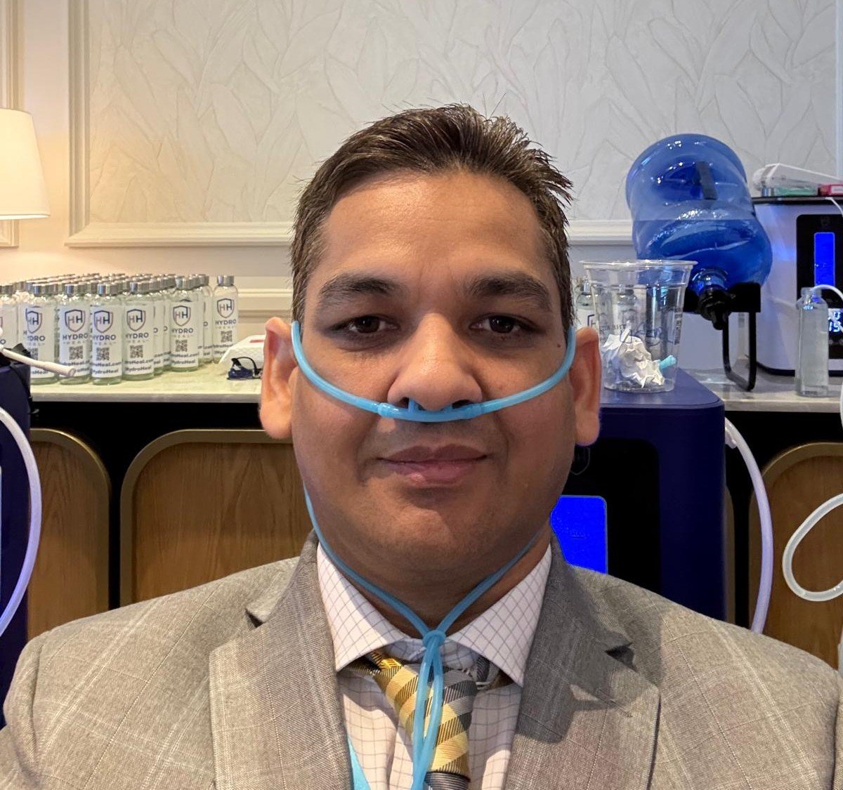 Man in a suit with a nasal oxygen tube, sitting in a room with medical and water equipment behind him.