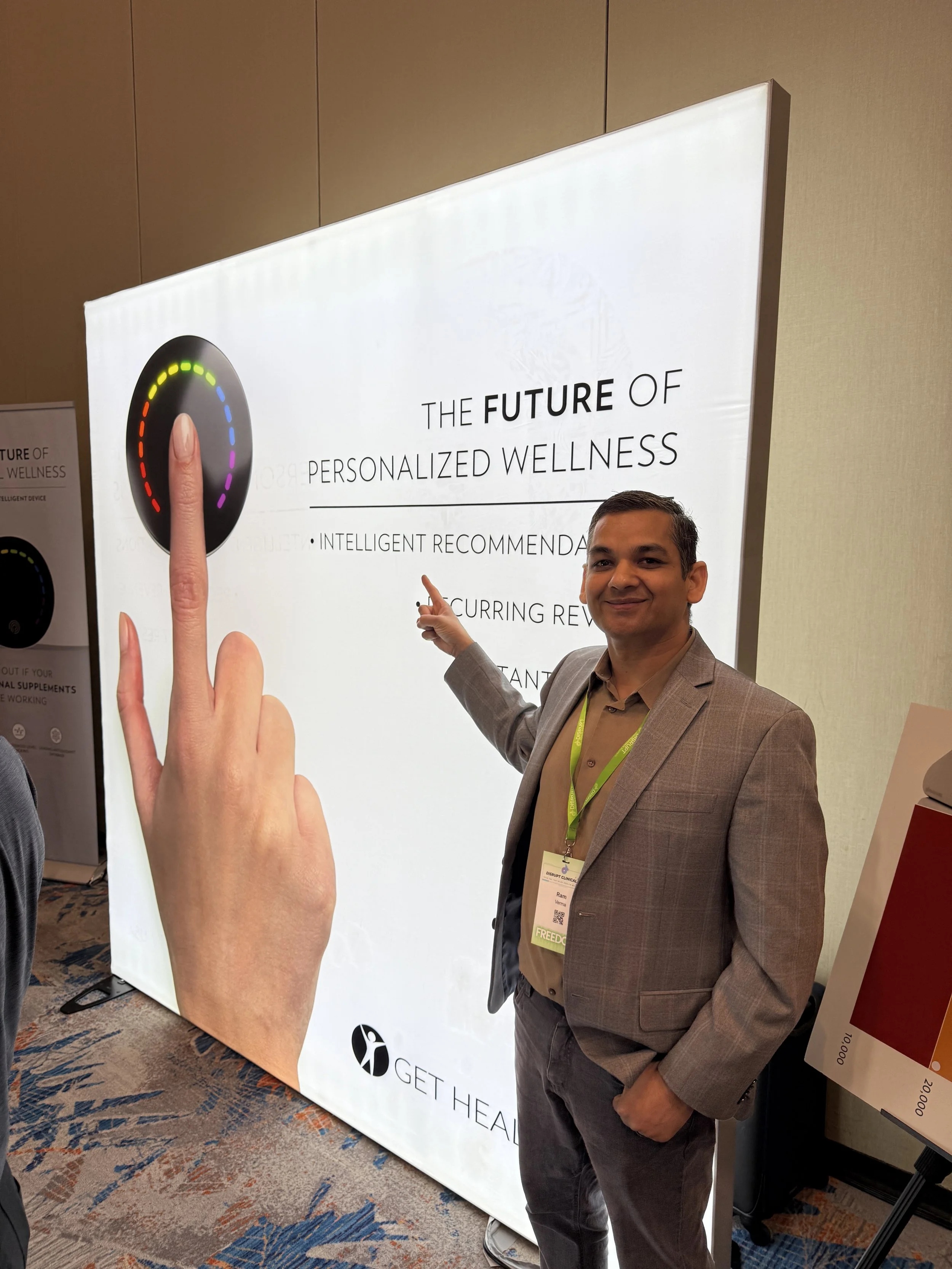A man in a gray suit and an orange shirt standing in front of a large white display board with text about personalized wellness. The man is smiling and pointing at the board.