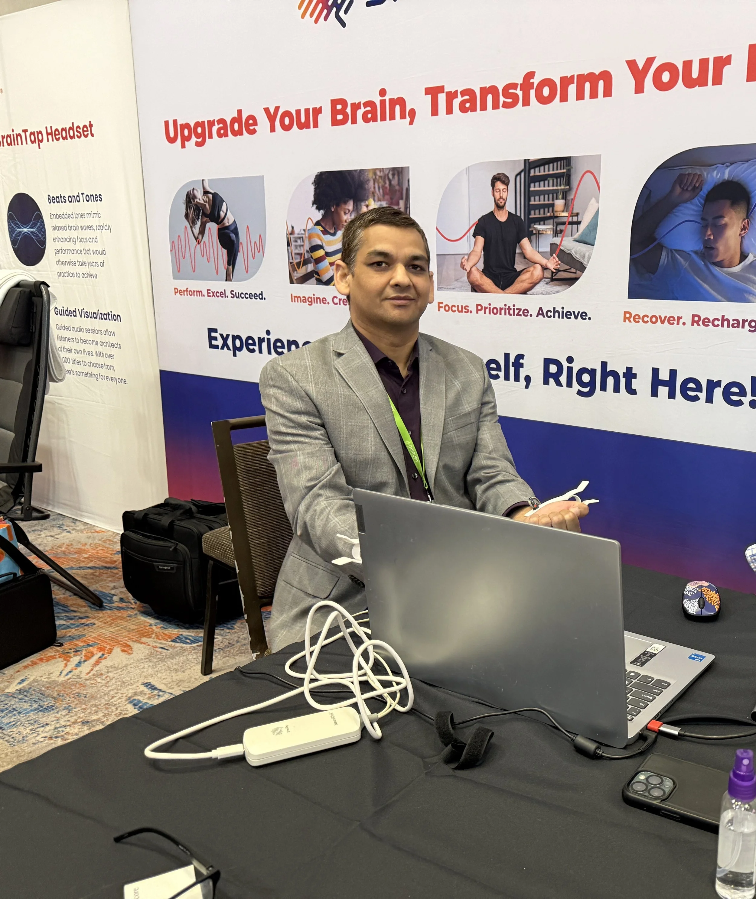 A man in a gray blazer and dark shirt sitting at a table with a laptop and a smartphone, with a large promotional poster in the background advertising brain training. The poster features images of people practicing mindfulness and meditation, with text about upgrading the brain, transforming, focusing, and achieving.