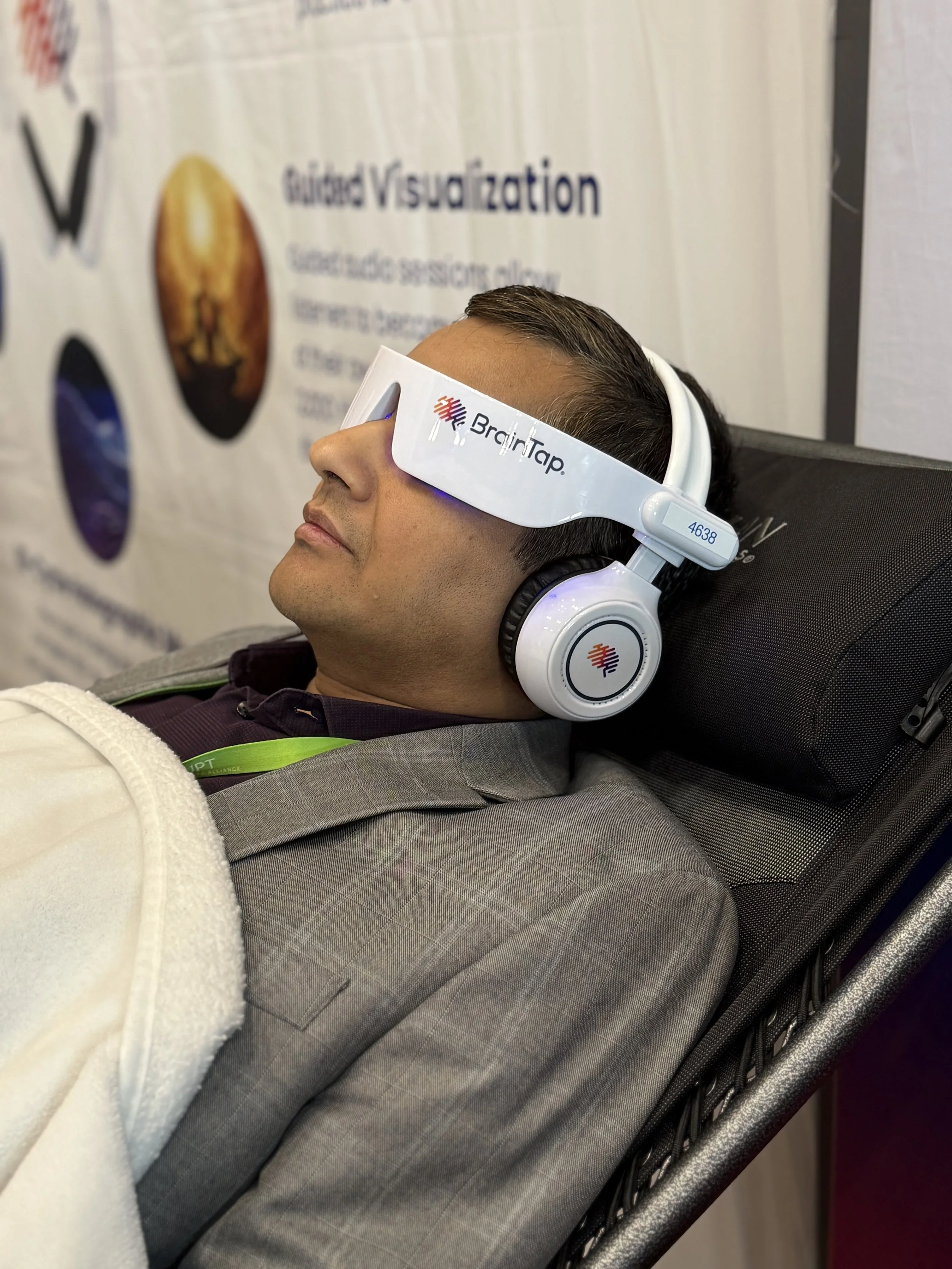 A man wearing white BrainTap headphones resting on a black chair with a white towel across his chest, in front of a poster that reads 'Guided Visualization'.