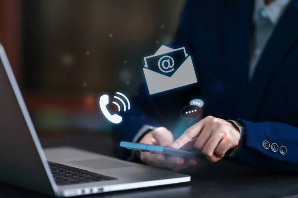 Person in business attire using a smartphone with digital icons of email, phone, and chat overlaid, in front of a laptop.