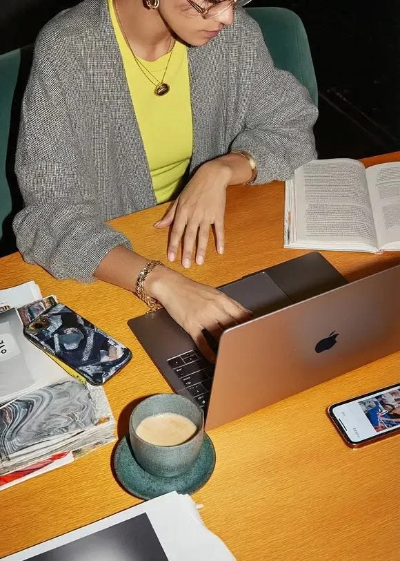 Woman working on a laptop at a cluttered wooden table, with a mug of coffee, papers, a smartphone, and an open book nearby.