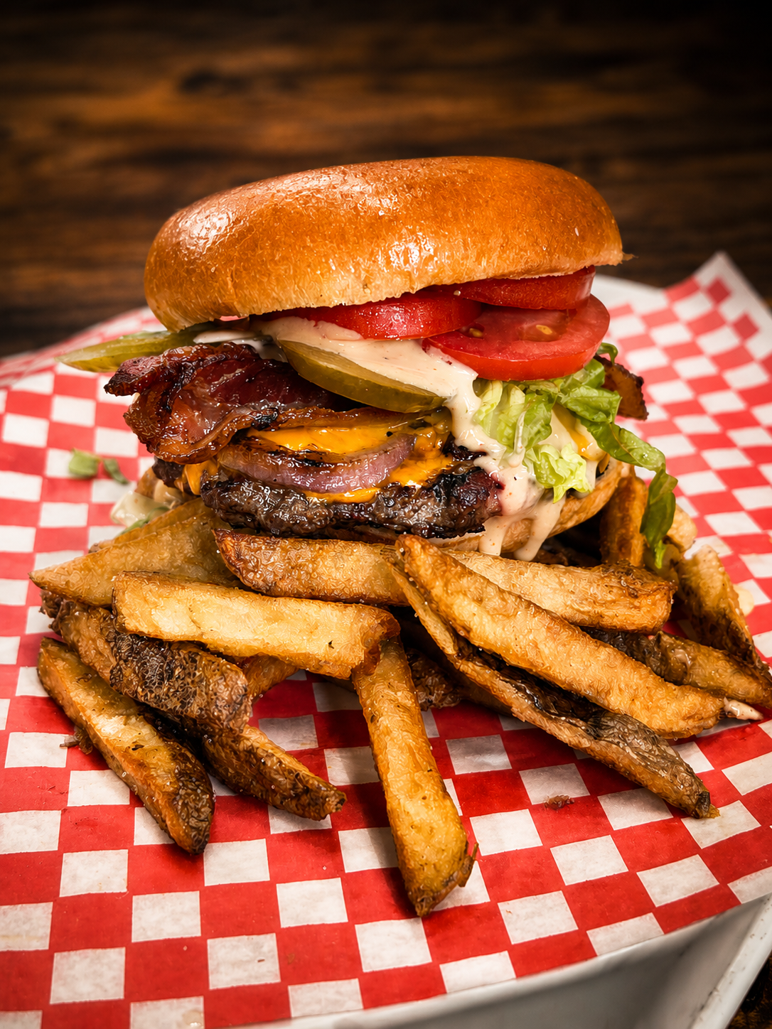 A cheeseburger with bacon, pickles, lettuce, tomato, onion, and mayonnaise on a bun, served with seasoned French fries on red and white checkered paper.